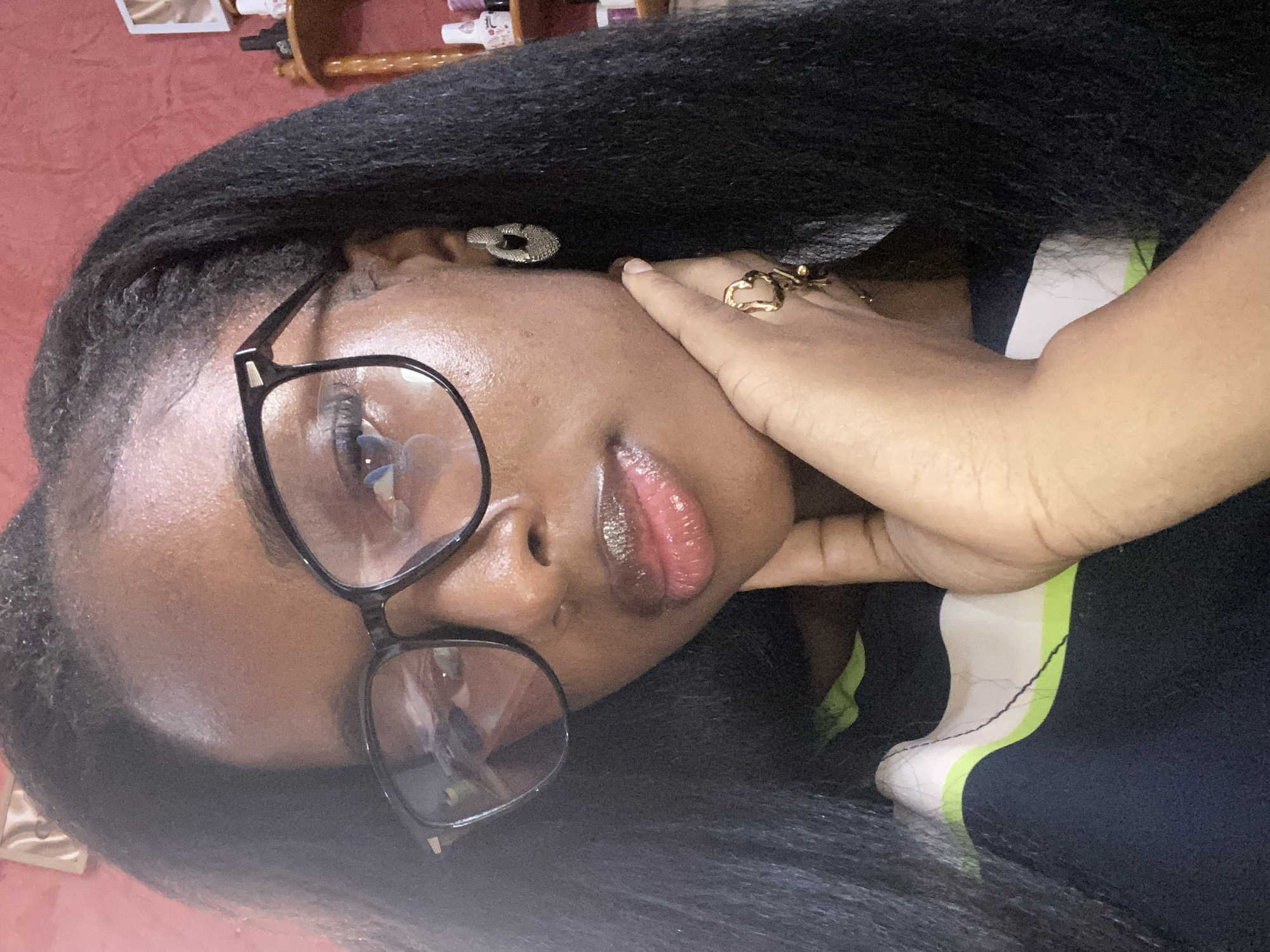 A close-up, selfie-style photograph shows a Black woman wearing large, black-framed eyeglasses. She has dark, long, textured hair, which is styled to the sides. Her lips are glossy, and she is wearing lip gloss. A silver, textured hoop earring is visible in her right ear, and a gold ring with a heart design is on her left hand, which is resting on her chin. She is wearing a dark-colored top with a stripe of white and light green. The background is a textured, reddish wall, with a shelf of bottles visible to the right. The image is taken indoors, likely in a residential setting within Jos, Nigeria.