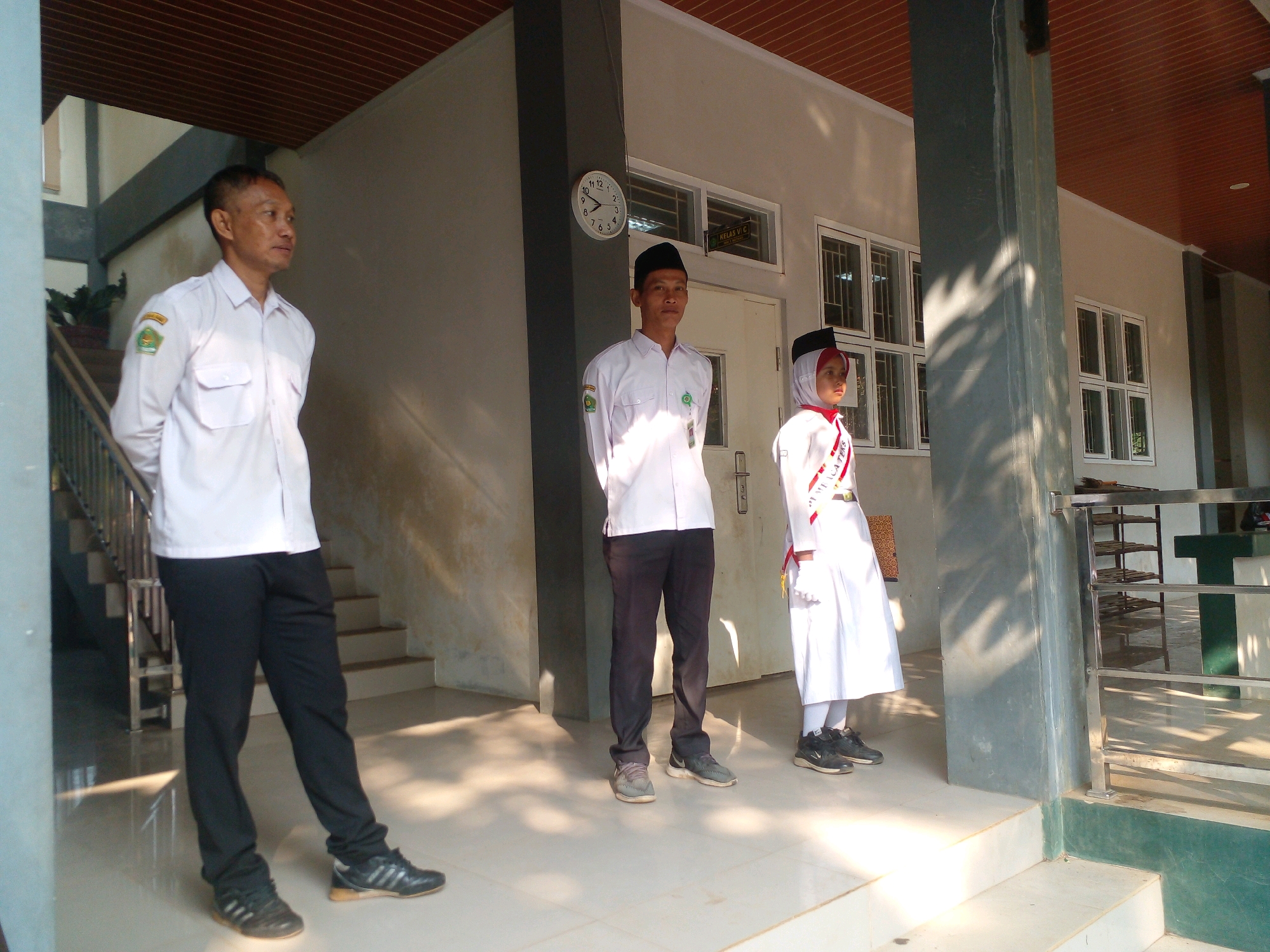 In the city of Banyuputih, Indonesia, three individuals are standing on a tiled outdoor walkway in front of a building with a white facade and wooden soffits. The building has multiple windows, some with bars, and a prominent clock on the exterior wall. To the left, a man in a white collared shirt with patches and black pants stands facing right, his hands clasped behind his back. To his right, a young male in a similar white shirt and black pants, wearing a black peci (a traditional cap), looks forward. Further to the right, a young female in a white uniform, including a headscarf and a red and white sash, stands facing left. Behind them, a set of stairs with metal railings leads up. The scene is illuminated by sunlight, casting shadows on the ground.