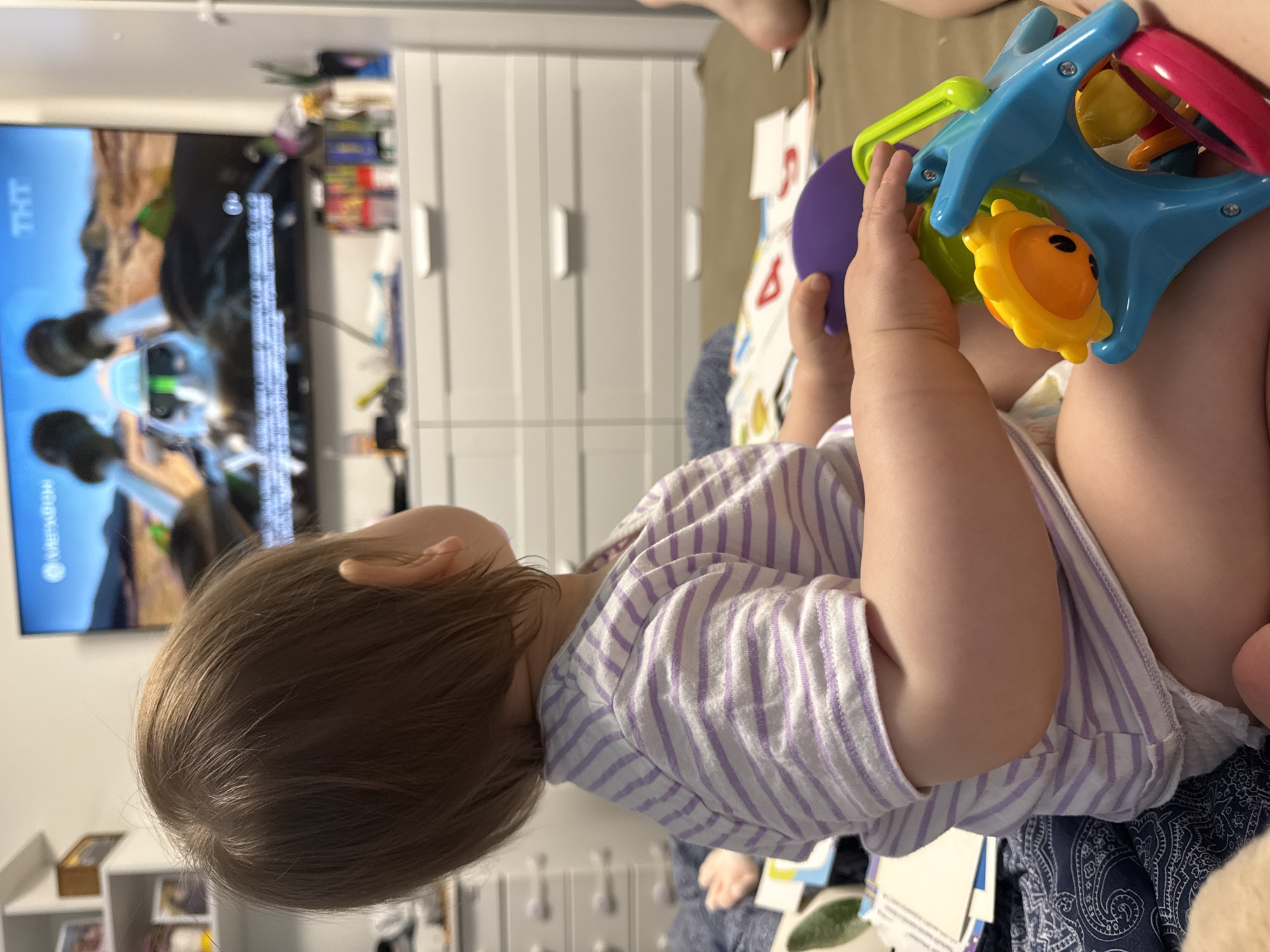 A baby is sitting and holding a colorful toy. The baby is wearing a white and purple striped shirt and a diaper. A television is visible in the background. The television is turned on and shows a landscape. A white dresser is also visible in the background. The location is not discernible from the image.