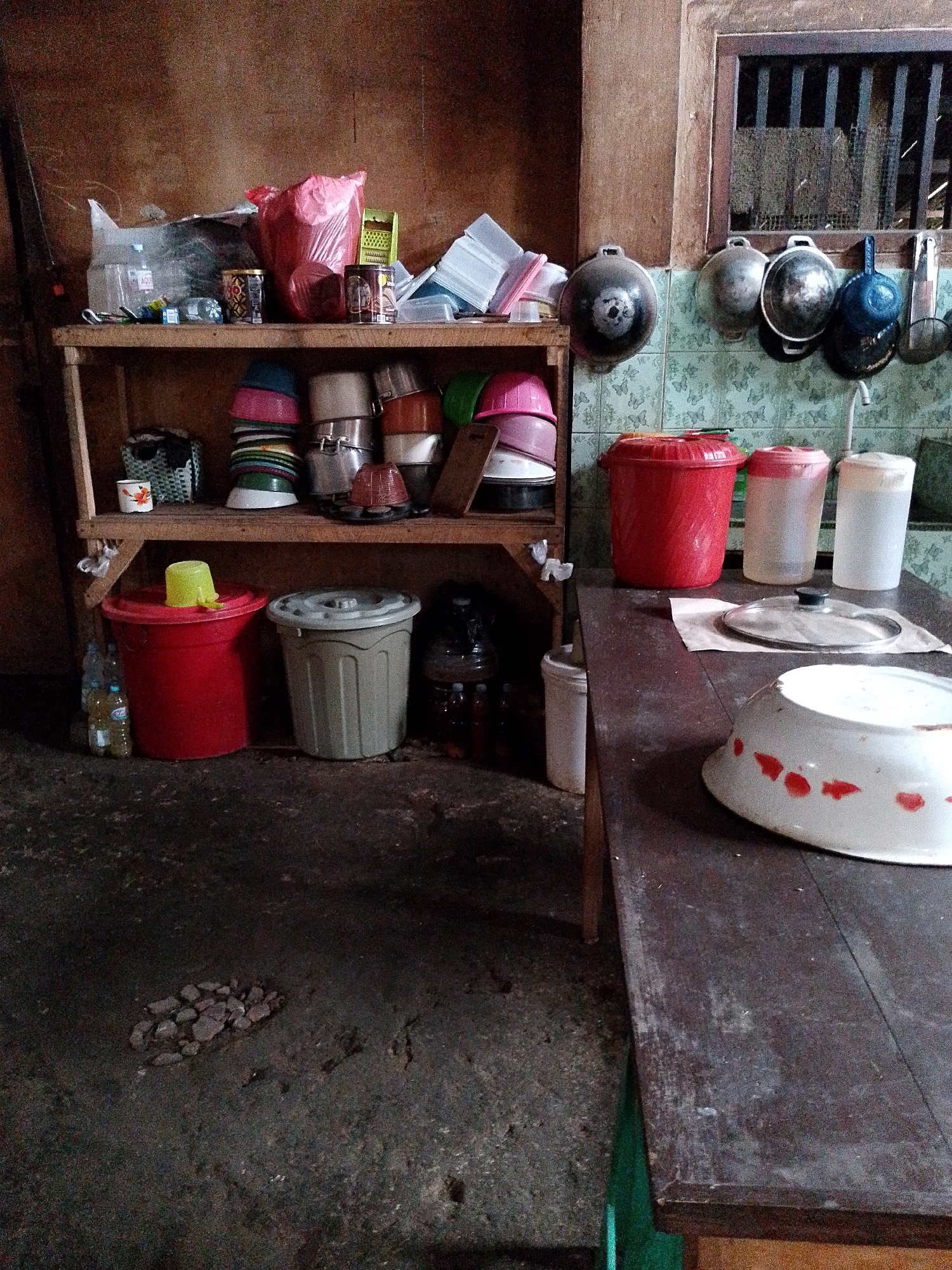 The image depicts a cluttered kitchen area. A wooden shelf unit stands against a wall. The top shelf holds a red plastic bag, some containers, and other miscellaneous items. The shelf below contains stacks of bowls and pots. In the foreground, on the left, are two large plastic bins, one red and one grey, with a few bottles of liquid next to them. To the right, a wooden table is visible, with a glass lid and a large white bowl with red markings. A tiled wall with a barred window can be seen in the background. The floor appears to be concrete. This scene is located in a kitchen in Babadan, Indonesia, likely a residential setting. There are no people or animals visible in the image.