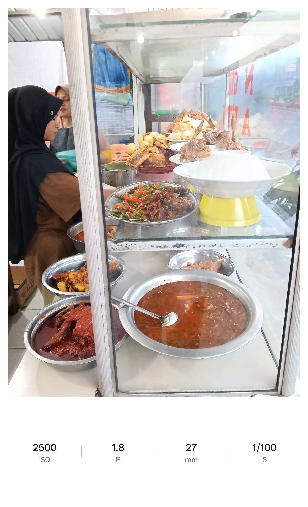 A well-lit, medium shot depicts a food stall in Lhokseumawe, Indonesia. The stall is enclosed in glass, displaying various cooked dishes in metal bowls. On the bottom shelf, a large bowl contains a dark red sauce with visible pieces of meat, while another adjacent bowl has a lighter brown sauce. The middle shelf features fish dishes with vegetables, and a large white bowl of rice sits on a yellow stand. The top shelf contains fried foods and more fish dishes. A woman wearing a black hijab and brown dress is visible on the left, seemingly interacting with the stall. The environment appears to be urban, with some street details reflected in the glass.