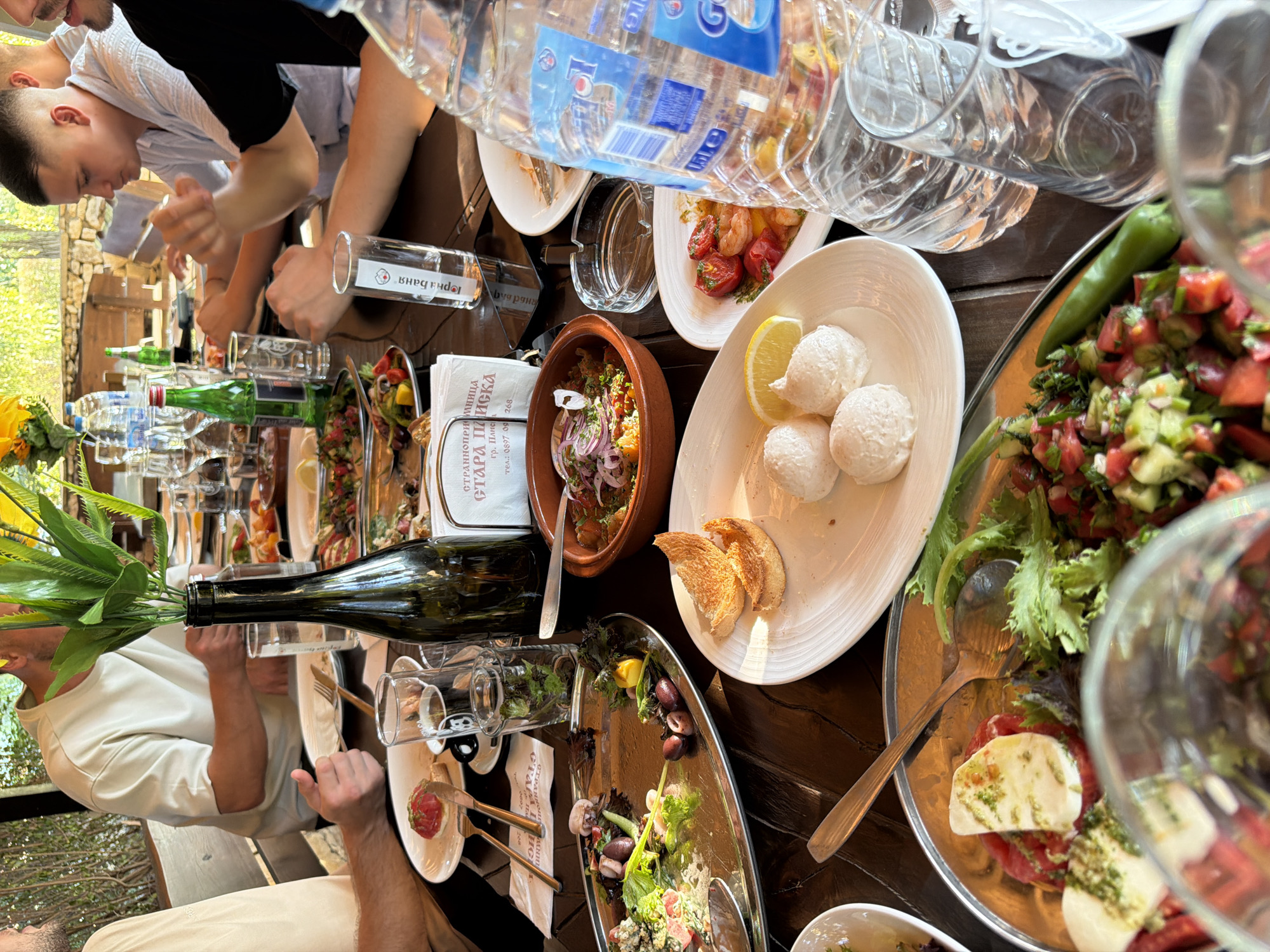 An overhead, close-up shot depicts a gathering of people enjoying a meal at an outdoor restaurant, specifically identified by a nearby sign as "STRANO PRIYATELSTVO TARA PANIČKA" in Pliska, Bulgaria. The table is laden with a variety of dishes, including salads, appetizers, and what appears to be a stew. Empty and partially consumed plates, glasses, cutlery, and bottles of water and wine are scattered across the wooden surface. Two men are visible in the background, engaged in eating. The atmosphere suggests a casual and convivial dining experience. The setting includes foliage and sunlight filtering through trees, indicating it's daytime.