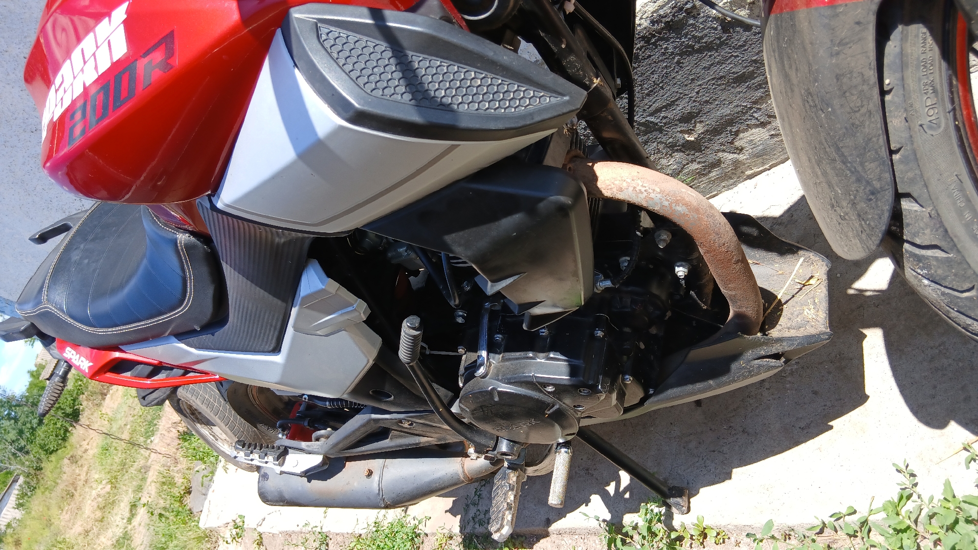 The image shows a close-up, top-down view of a red and silver motorcycle parked outdoors on a concrete surface, with some dry vegetation and grass visible around it. The motorcycle's engine, exhaust pipe, footpegs, and part of the seat and fuel tank are visible. The fuel tank has white lettering that appears to read "200R." The scene is illuminated by natural daylight, casting shadows on the ground. The background consists of a grassy incline with trees and a hint of a building. There are no people or animals present.
