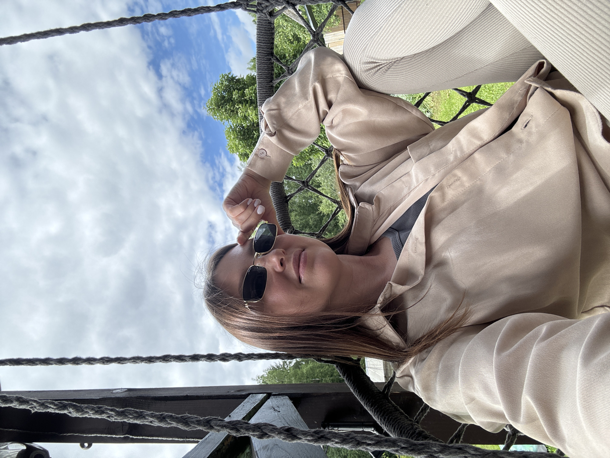 A young woman with long, brown hair wears sunglasses, a tan button-up shirt, and cream-colored pants. She is sitting in a rope swing, with thick ropes on either side of the frame, and a dark wooden structure behind her. Green trees and a blue sky with white clouds are visible in the background. The woman is leaning her head on her hand, looking to the side. The image appears to be taken outdoors in the city of Ponazyrevo, Russia.