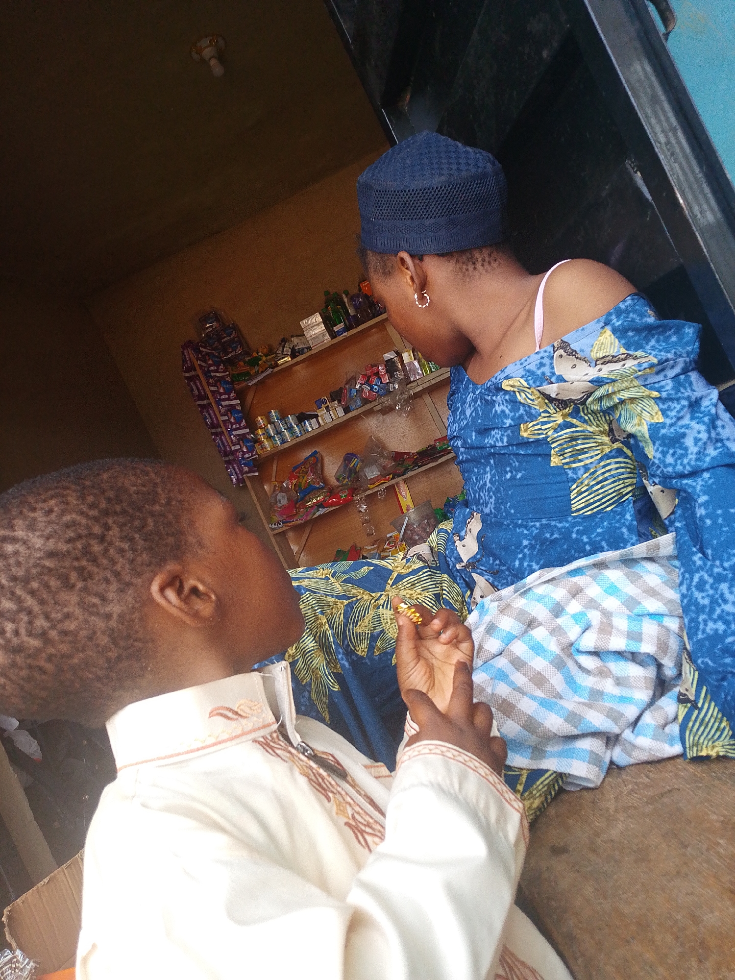 The image depicts an indoor scene, possibly a small shop or home, in Abeere, Nigeria. In the foreground, a young child is seated, facing away from the camera and to the right. The child is wearing a cream-colored garment with embroidered detailing around the collar and down the front. The child's right hand is raised, and it appears to be holding a small, golden object.
To the right of the child, and slightly behind, is an adult woman, also seated. She is wearing a dark blue, textured cap on her head. Her visible shoulder is bare, and she is wearing a blue garment adorned with a yellow and white floral pattern. She is looking down and away from the camera. A pink bra strap is visible on her shoulder. Her left hand is resting on her lap, and she is wearing a gold hoop earring in her visible left ear.
In the background, shelves are stocked with various items, including bottled liquids, packaged goods, and what appear to be snacks or confectionery. The shelves are made of wood, and the wall behind them is a warm, light brown color. The lighting in the scene is somewhat dim, suggesting it might be late afternoon or that the interior is not brightly lit. The overall impression is a candid moment captured within a modest commercial or domestic space.