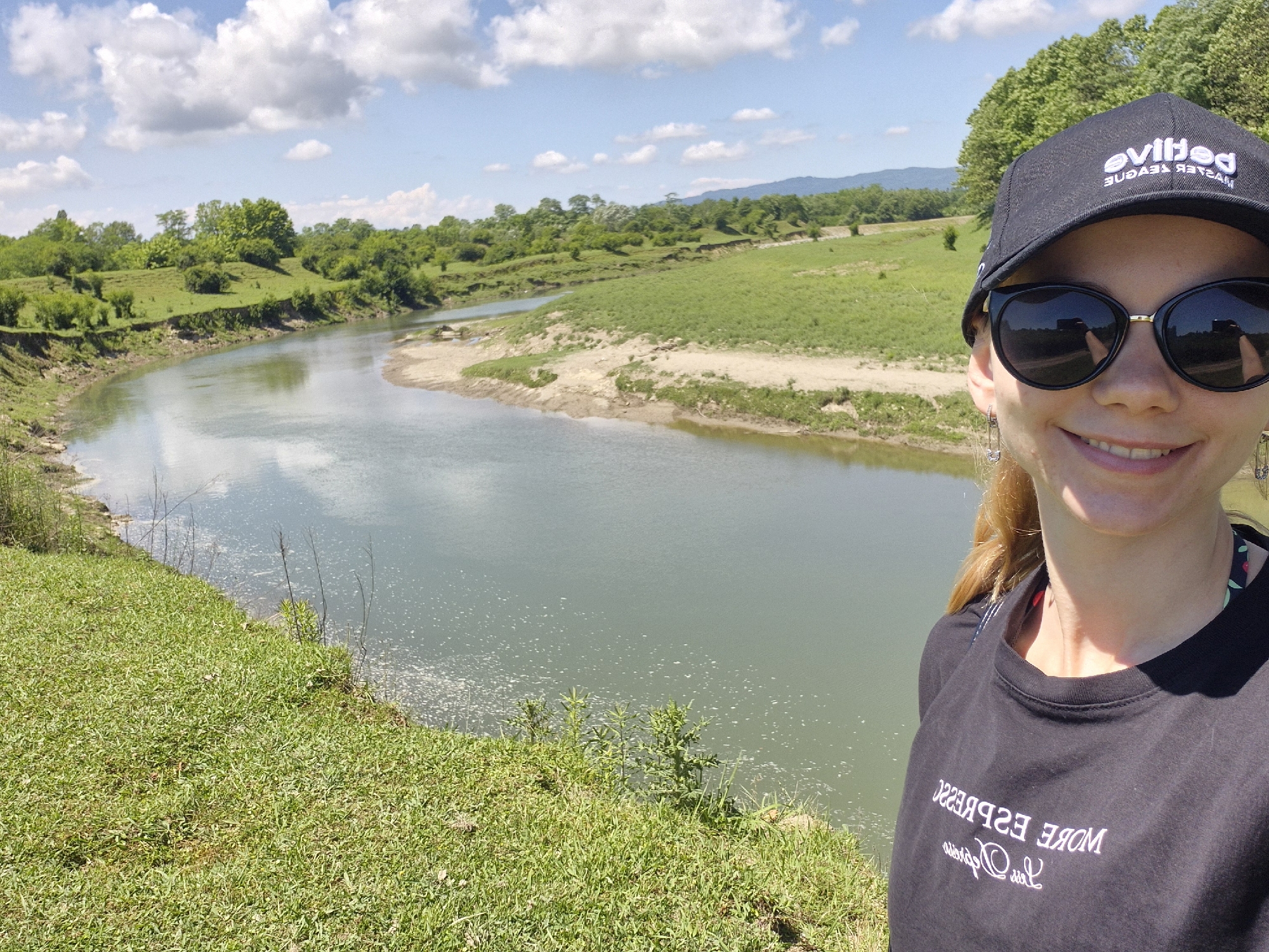 A woman wearing sunglasses and a black baseball cap takes a selfie in front of a river. The river is wide and calm, with a sandy bank on one side and grassy hills on the other. Trees and vegetation line the riverbanks, and mountains are visible in the distance. The woman is smiling and her hair is pulled back. She is wearing a black t-shirt. The scene appears to be a natural landscape, likely in a rural area. The location is identified as Darcheli, Georgia.