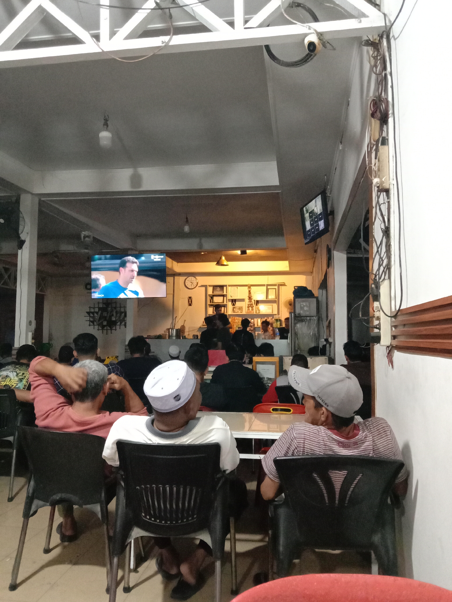 The image depicts an indoor gathering, likely a cafe or restaurant, with several individuals watching a television screen. The television, mounted high on a wall, displays a man in athletic attire. The room has a simple, utilitarian aesthetic, with exposed ceiling beams and industrial-style lighting. In the foreground, three men are seated facing away from the camera, their attention directed towards the television. One man wears a white peci, a traditional cap often worn by Muslim men. Another man wears a grey baseball cap. The atmosphere appears relaxed, with patrons casually observing the broadcast. The presence of what appears to be a CCTV camera mounted on the ceiling suggests a security-conscious environment. The overall impression is of a communal viewing experience, possibly for a sporting event or other popular program. The context suggests the location is a public establishment in Lhokseumawe, Indonesia, where such gatherings are common.