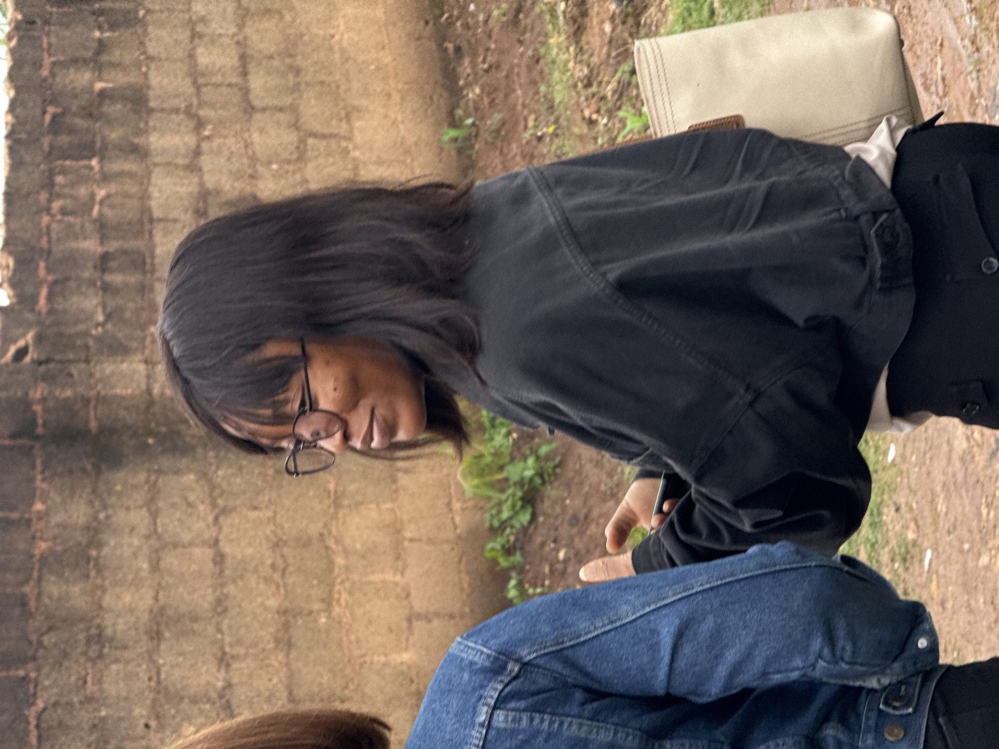 The image shows two individuals from the torso down, sitting outdoors. The person in the foreground, wearing a blue denim jacket, is partially visible, with their legs crossed. The second person, to the left and slightly behind, is wearing a black jacket and glasses. They are looking downwards, with their face partially obscured by their hair. The background suggests an outdoor setting with a brick wall to the left and a dirt path with sparse vegetation to the right. A light-colored bag is placed behind the person in the black jacket. The lighting suggests it is daytime.