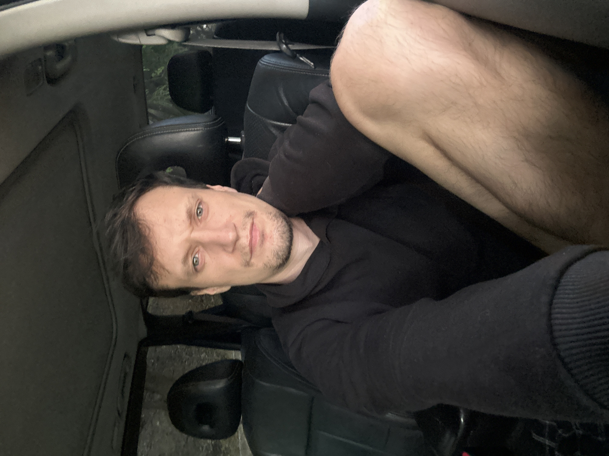 A man is sitting in the driver's seat of a car, possibly parked on a road in Makiv, Ukraine. He is looking directly at the camera, his expression neutral. The interior of the car is visible, with black leather seats and a sunroof. The lighting suggests it is daytime, but the mood is somewhat dim. The man is wearing a black hooded sweatshirt. His bare leg is visible in the foreground, resting on the seat. The background outside the car appears to be wooded.