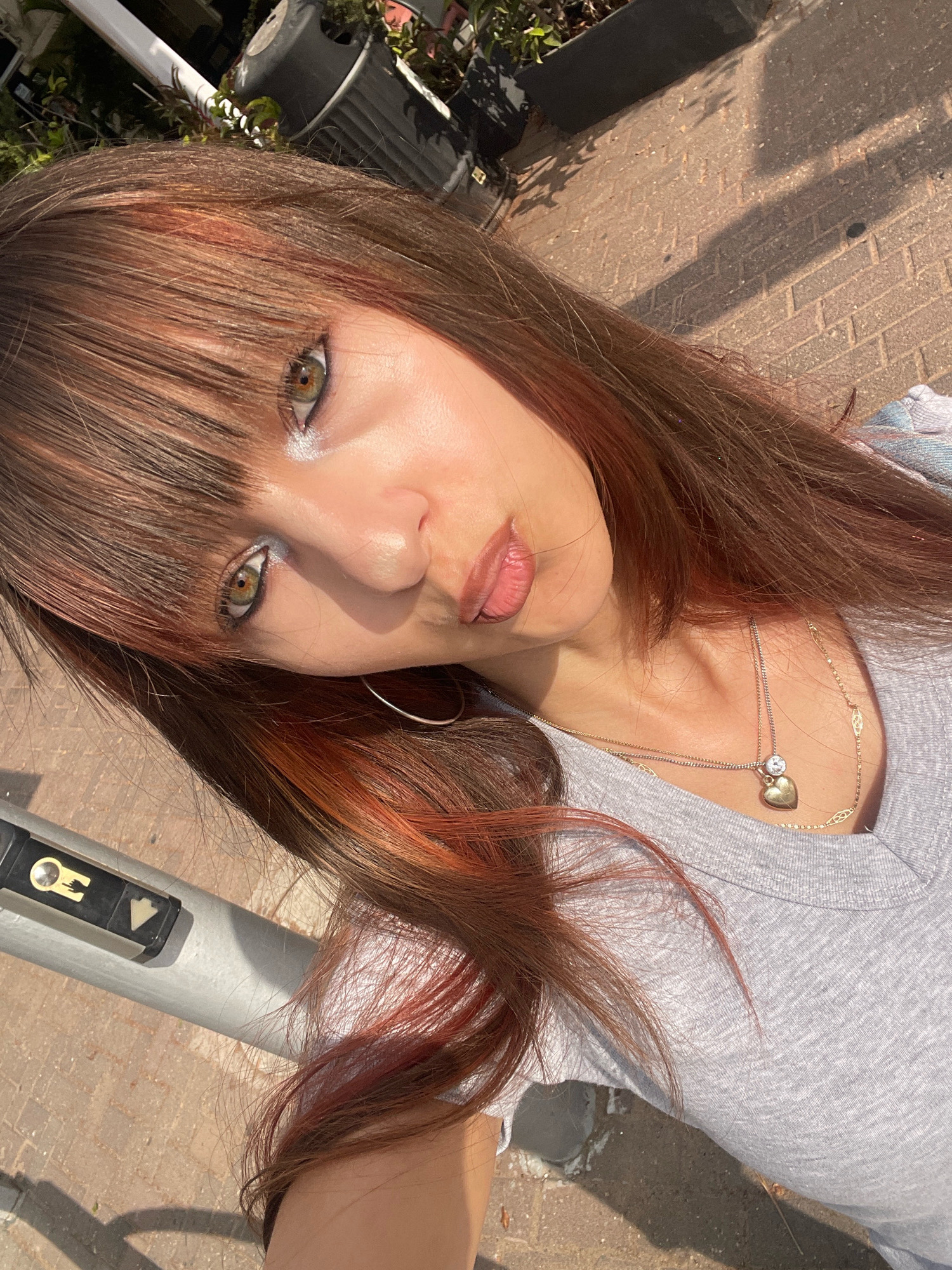 A young woman with long, brown hair with red highlights is pictured from the chest up, tilted at an angle, in an outdoor urban setting. She is wearing a grey t-shirt and a necklace with a heart-shaped pendant. Her makeup includes eyeshadow and lip gloss. In the background, a brick-paved ground is visible, with shadows cast by sunlight. A metal pole with a button for pedestrian crossing is to her left. The scene appears to be in Raanana, Israel.