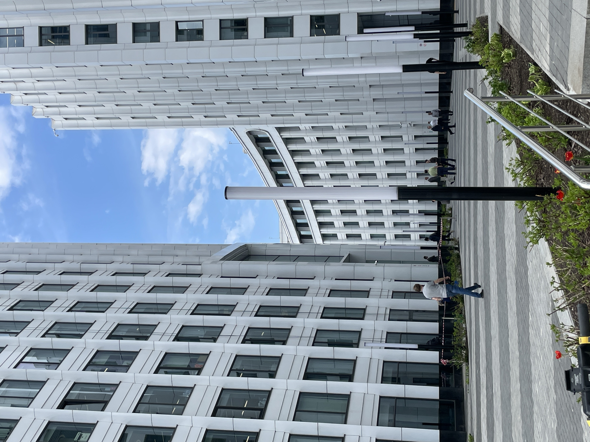 The image shows an urban scene between two tall, modern buildings with a clear blue sky visible between them. The buildings are characterized by a repetitive grid of windows and a white or light gray facade. A pedestrian walkway separates the buildings, paved with alternating light and dark gray stripes. Several people are walking along the walkway, some appearing to be individuals and others in small groups. A man in a light-colored shirt and blue jeans is walking on the right side of the image. Vegetation lines the edge of the walkway, with some greenery and a few red flowers visible. Modern light fixtures or poles are positioned along the walkway. The perspective of the image is slightly tilted. The location in the city of Unknown, Unknown cannot be determined.