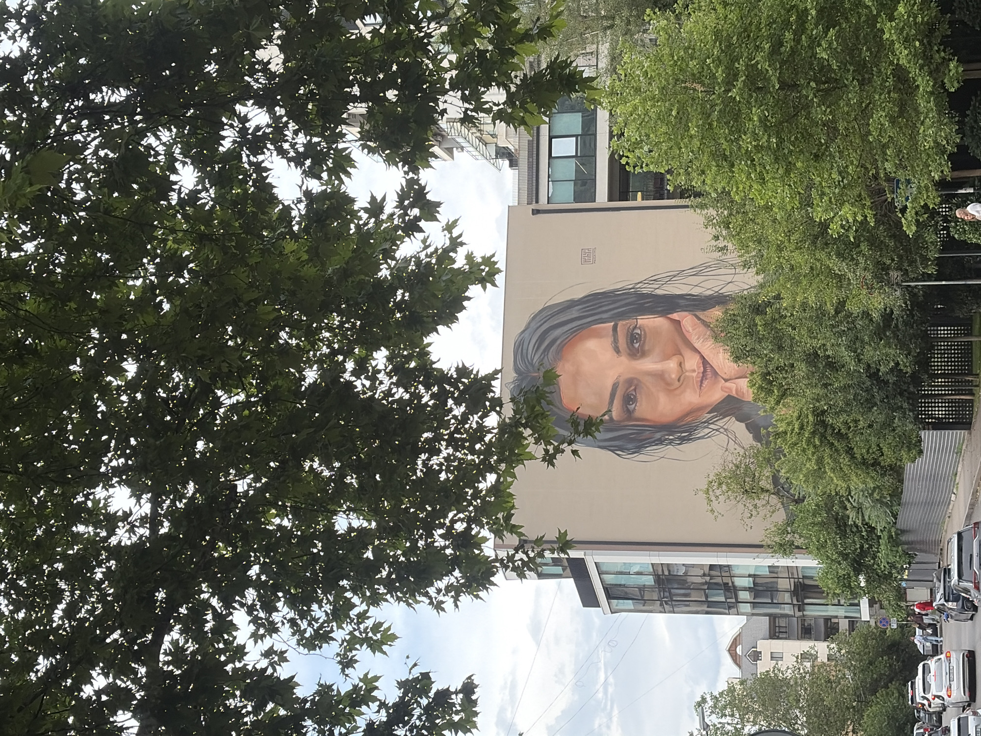 A large mural of a woman's face is painted on the side of a modern building in Tbilisi, Georgia. The woman's face is the dominant feature of the mural, with her eyes looking directly at the viewer. She is resting her chin on her hand, and her hair is wet and falling around her face. The building itself is made of glass and concrete, and it appears to be an office building or apartment complex. In front of the building, there is a street with cars and trees. The sky is overcast. The mural is located on the street side of the building, making it visible to people passing by.