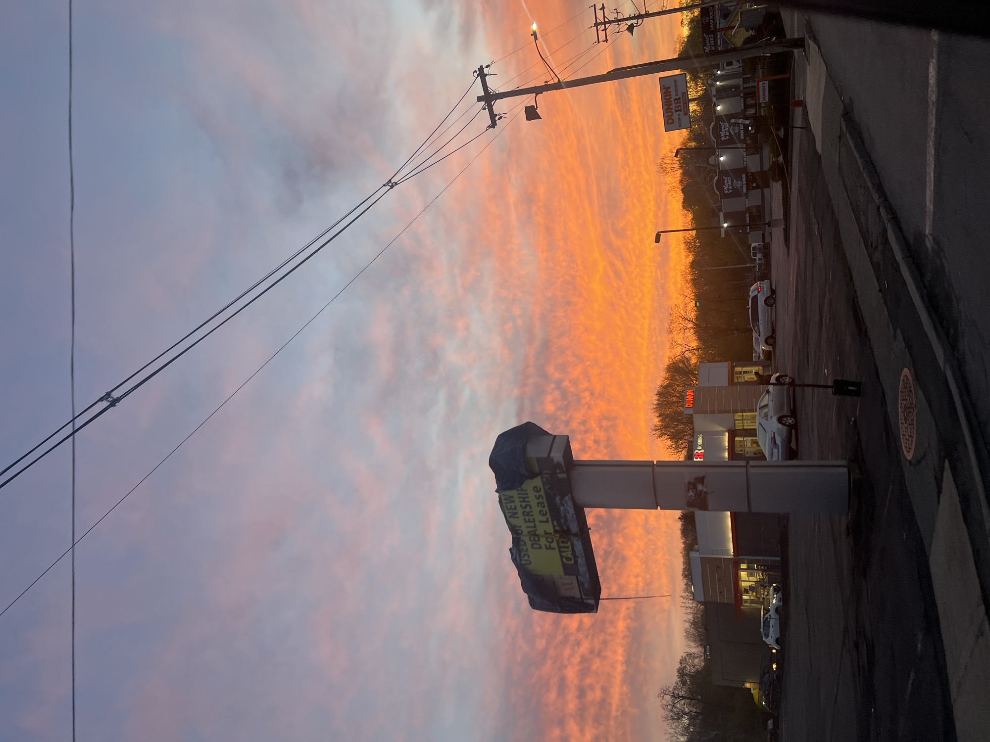 Here's a concise description of the image:
The image captures a vivid sunrise or sunset in a commercial area, possibly in the City of Newburgh, United States of America (based on the identified business names). The sky is filled with streaks of orange and pink clouds. Several signs are visible, including one advertising a "USED OF NEW DEALERSHIP For Lease" and another for "DUNKIN' BASKIN BR ROBBINS". Several other businesses and parking lot can be seen, the image appears to be a perspective from a roadside, utility lines and poles are also featured in the sky.