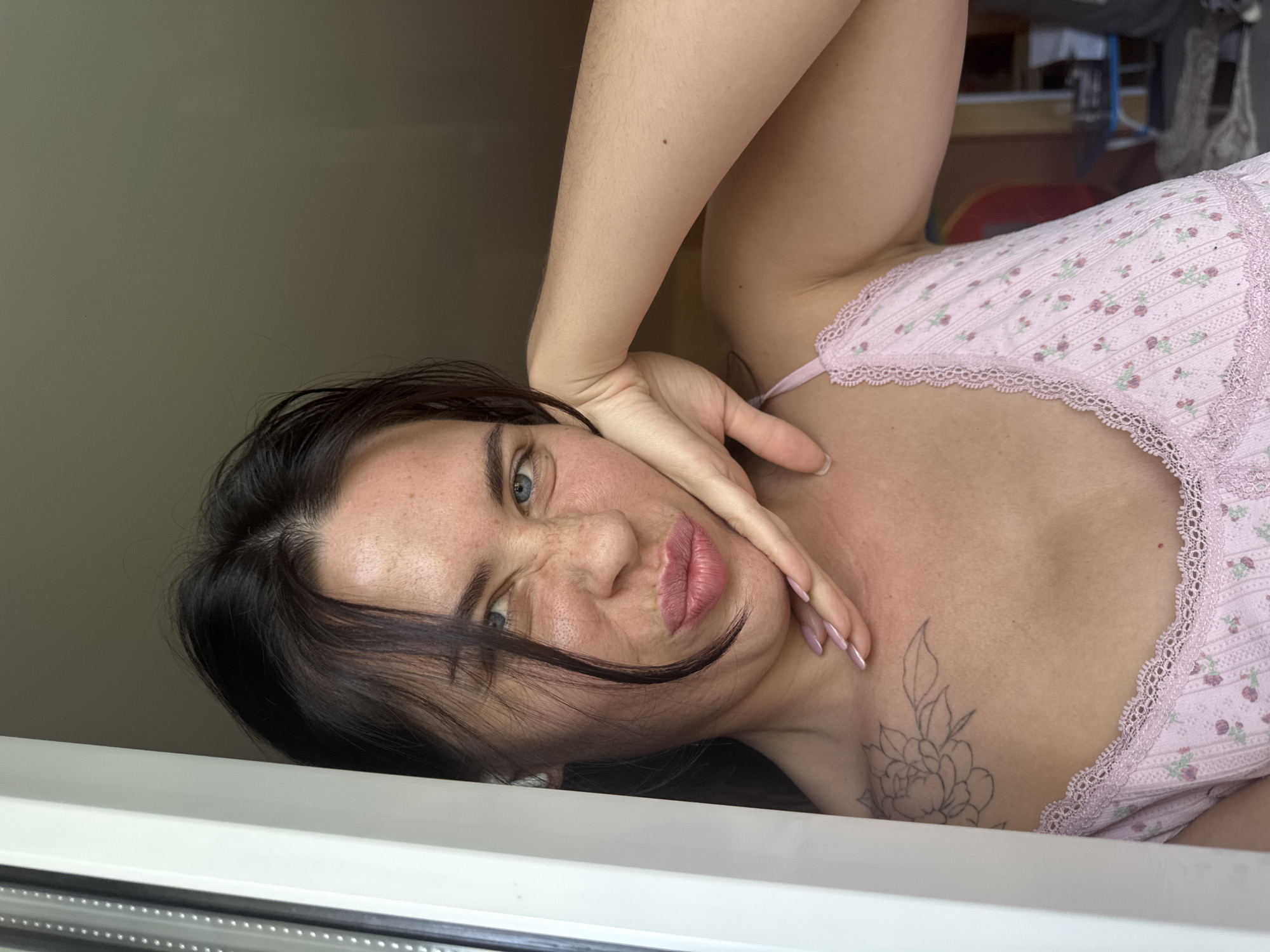 A young woman with dark hair and blue eyes is pictured close-up, leaning against a window frame. She is wearing a pink floral camisole. Her face is turned towards the camera, and she is making a puckered expression with her lips. Her right hand is placed on her cheek, and her left arm is bent, resting on her chest. A tattoo of a flower is visible on her left shoulder. The background appears to be an indoor setting, possibly a room with muted lighting. The image was taken in the city of Chaikovsky, Russia.