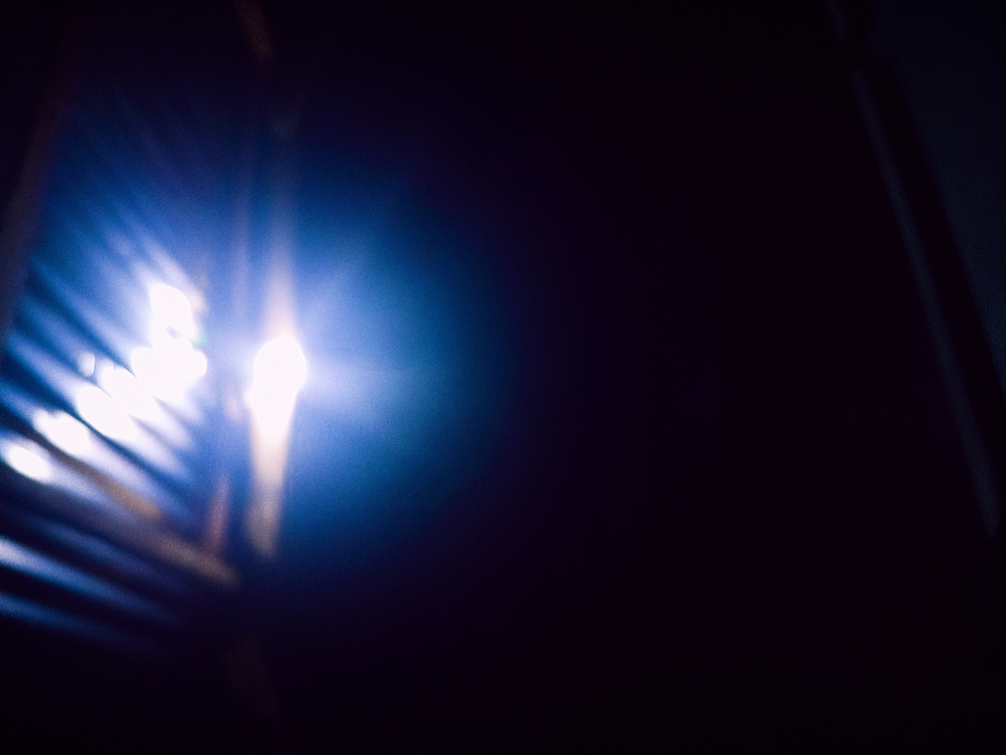 The image is a dark, abstract photograph featuring a bright, blown-out light source in the upper portion. The light appears to be coming from above and to the right, casting streaks and flares across the frame. There are some indistinct shapes visible in the background, possibly indicative of corrugated metal or a textured surface, illuminated by the intense light. The lower portion of the image is predominantly black, suggesting a deep shadow or unlit area. No specific location in Jalingo, Nigeria can be identified from this abstract image. There are no people, discernible objects, actions, or interactions visible.