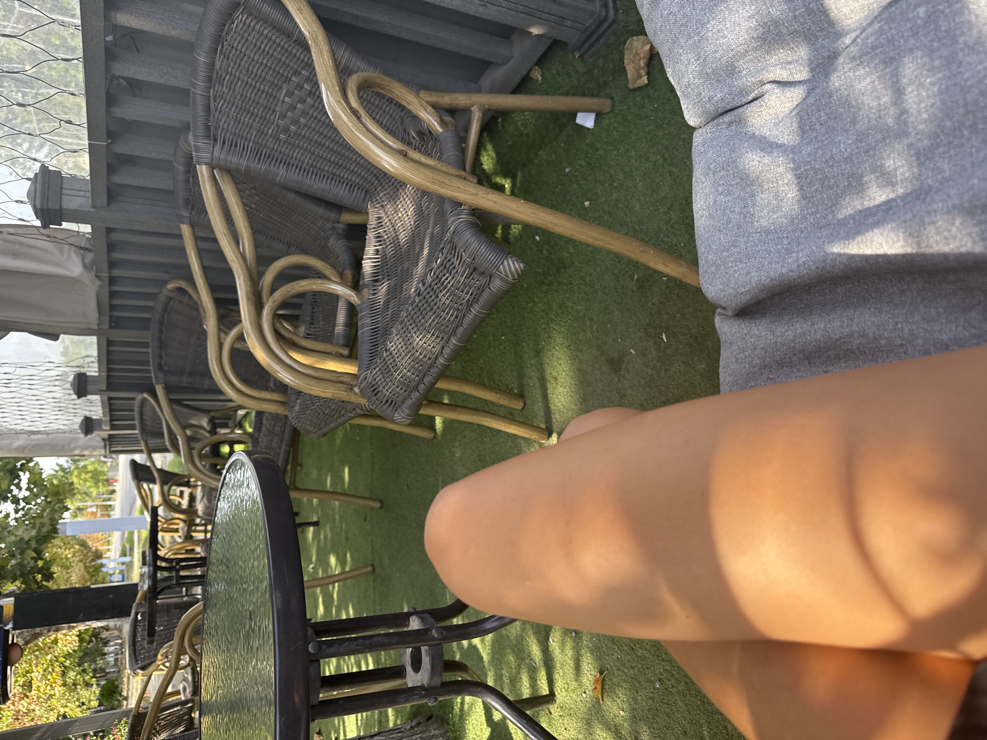 This image depicts an outdoor seating area, possibly at a cafe or restaurant. In the foreground, a person's bare legs and upper thighs are visible, illuminated by warm sunlight. The legs appear tanned. To the left, a round table with a dark, textured surface and metal legs stands on a green, artificial turf surface. Several outdoor chairs with dark wicker-like seating and curved wooden frames are arranged around tables in the middle ground. The chairs are empty. In the background, a dark railing with a lattice-like structure above it separates the seating area from an outdoor space that appears to be a street with trees and buildings. The overall scene suggests a casual, daytime setting. There is no specific indication or visual element in the image that places the location in Basarabeasca, Moldova.
