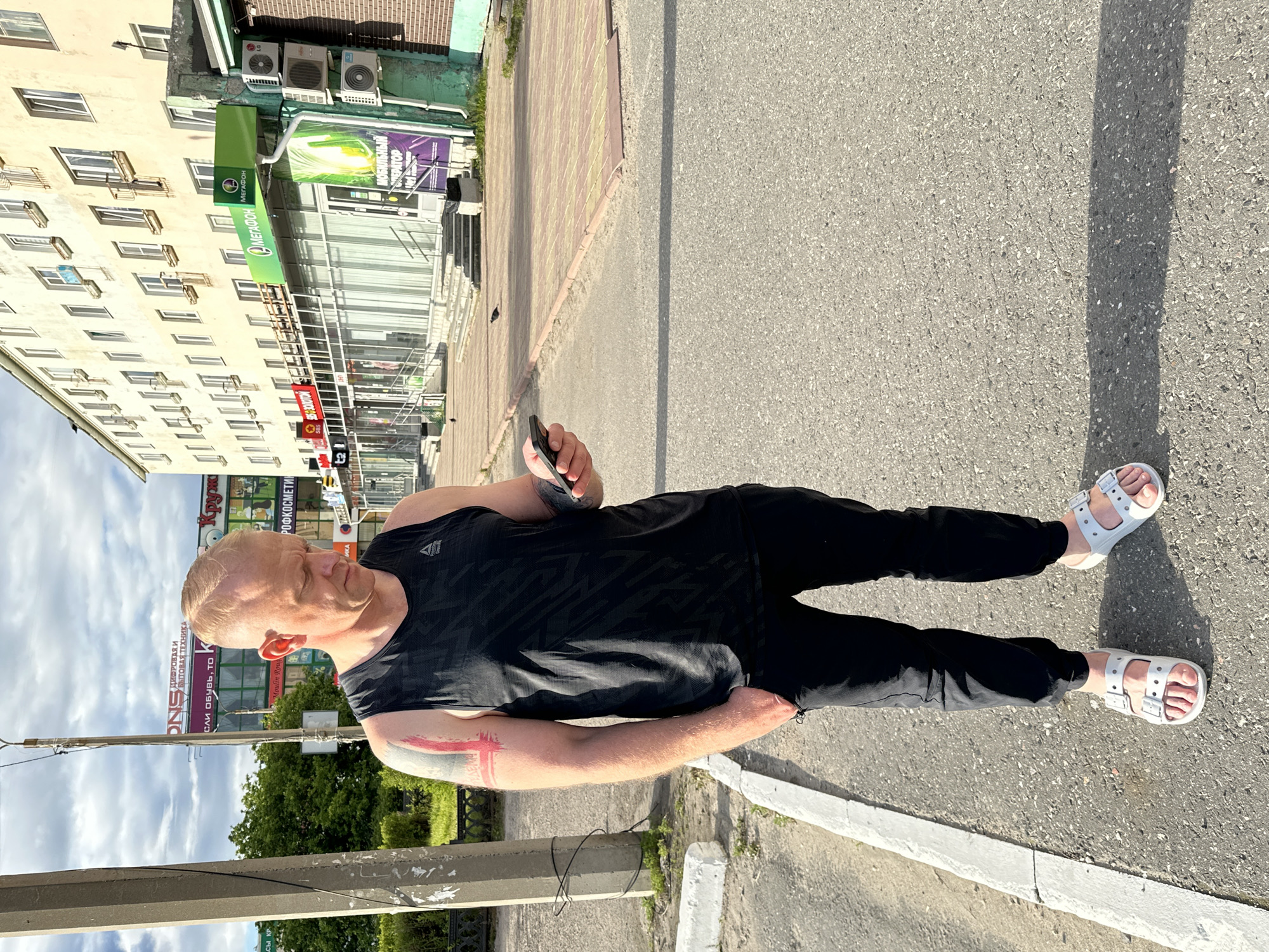 A bald white man wearing a black sleeveless Reebok t-shirt, black pants, and white sandals stands on a paved sidewalk in Monchegorsk, Russia. He is looking down at his phone, which he holds in his right hand. His left hand is in his pocket. He has a tattoo on his left arm. In the background, there is a light-colored apartment building with numerous windows and balconies. Several shops with signs in Russian are located on the ground floor of the building, including one with a green "MEGAFON" logo. The sky is partly cloudy. There are trees to the left of the apartment building.