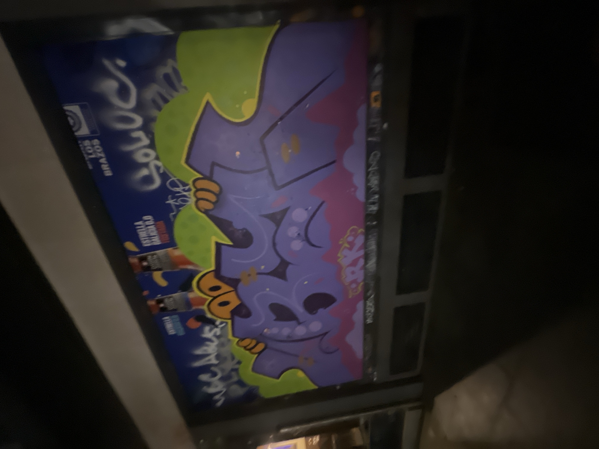 The image shows a graffiti mural on the exterior of a building in Barcelona, Spain. The mural features stylized lettering in purple with orange and green accents, depicting cartoon-like eyes and hands. The background of the mural is blue and includes text and logos, suggesting it may be an advertisement. The graffiti covers a large portion of the wall, which appears to be part of a storefront or commercial establishment. The scene is dimly lit, suggesting it was captured at night or in low light conditions. The surrounding environment is obscured by shadow, but appears to be an urban setting.