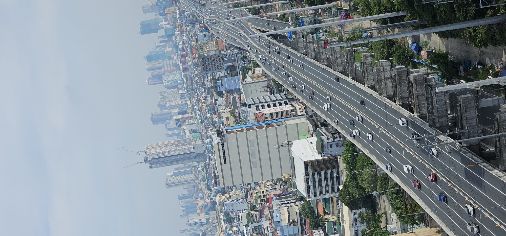 This is a high-angle, vertical, slightly tilted, daytime photograph of an elevated highway in Makati, Philippines. The highway, an intricate multi-lane structure with concrete support pillars, carries a moderate amount of traffic, consisting of cars, vans, and trucks. The road surface is grey asphalt with white lane markings. The highway curves and splits, creating a complex network.
In the foreground, the highway dominates the view, receding into the distance. On the left side of the highway, several buildings of varying heights and architectural styles are visible, including a large, light-colored rectangular building with horizontal dark bands. Further into the background, a dense urban cityscape unfolds, characterized by numerous high-rise buildings, including several under construction with cranes visible. The sky is overcast with a pale, hazy blue.
To the right of the highway, there are areas of green vegetation and more trees. Beneath the elevated sections of the highway, there are visible support structures and some makeshift shelters or storage areas with blue and orange tarps. The overall impression is of a busy, developed urban environment with ongoing infrastructure and construction.