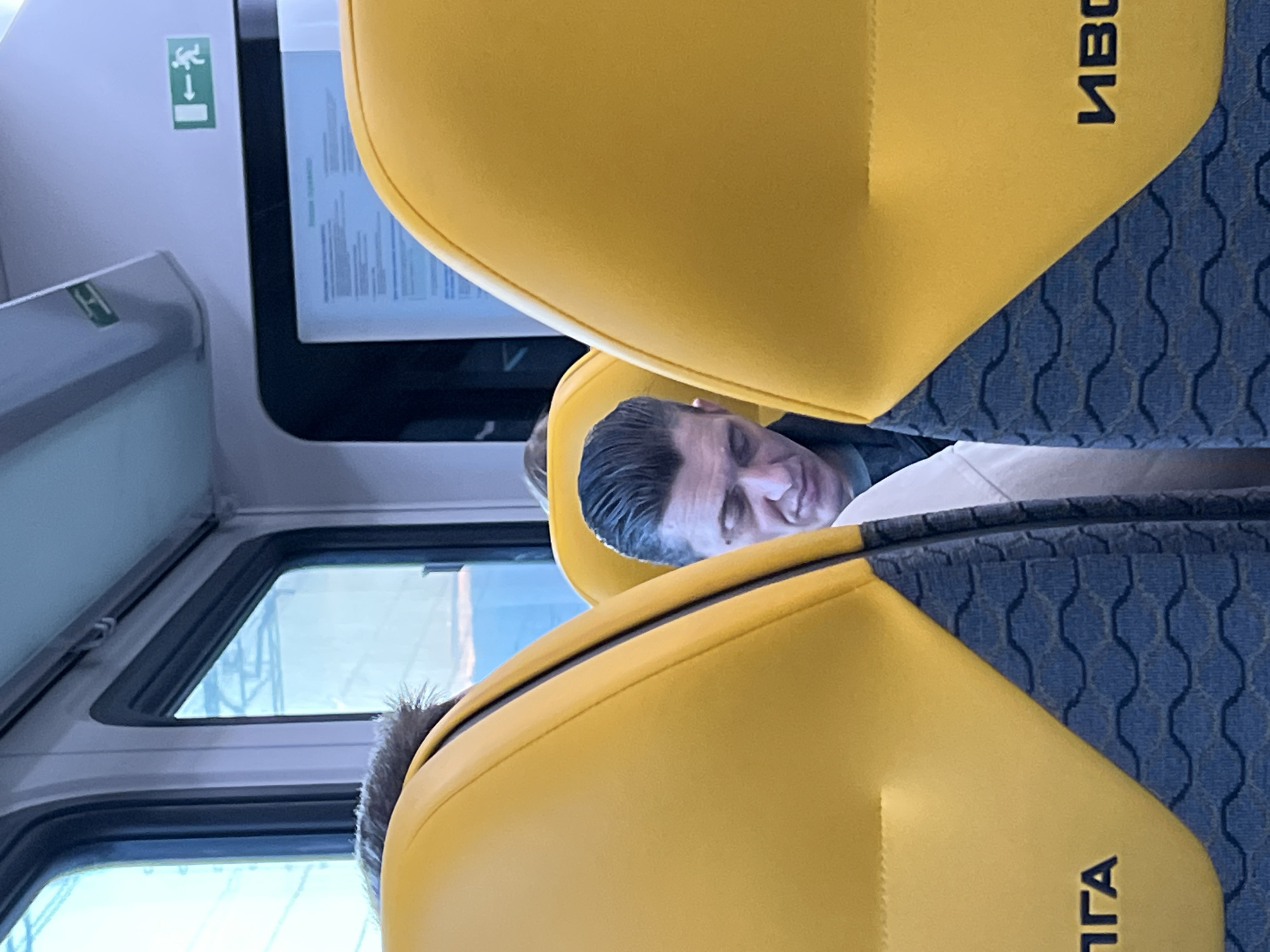 A man is sleeping on a train in Khimki, Russia. The train car is gray and white, with yellow and blue seats. An emergency exit sign is visible.