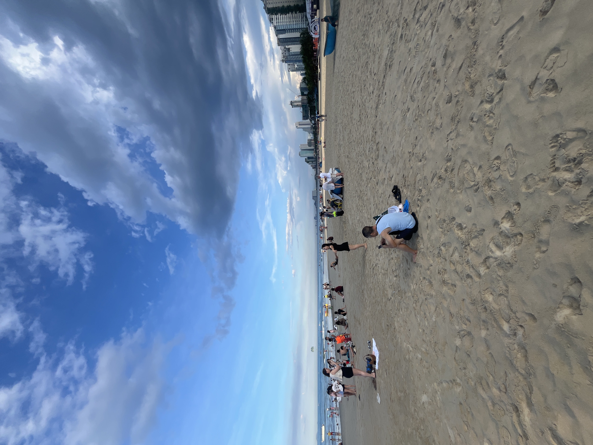 A panoramic view captures a busy beach scene along the coast of Da Nang, Vietnam. The sandy beach stretches wide, with numerous people scattered across it. In the foreground, a man in a light blue shirt sits on the sand, looking at his phone. To his left, a towel and flip-flops lie on the sand, with a blue bag next to them. Further into the mid-ground, a woman in a black dress stands facing left, appearing to talk to someone off-camera. To her left, a group of people are seated on the sand, including one wearing an orange life vest.
The beach extends to the water's edge, where people are wading and swimming. Small boats are visible in the distance on the ocean. Along the horizon, the skyline of Da Nang city is prominent, featuring numerous tall buildings and hotels. The sky above is a mix of bright blue and dramatic dark clouds, suggesting a dynamic weather system. Several blue fishing boats are present on the sand in the background. The overall atmosphere is lively, with many individuals enjoying the beach and the ocean.