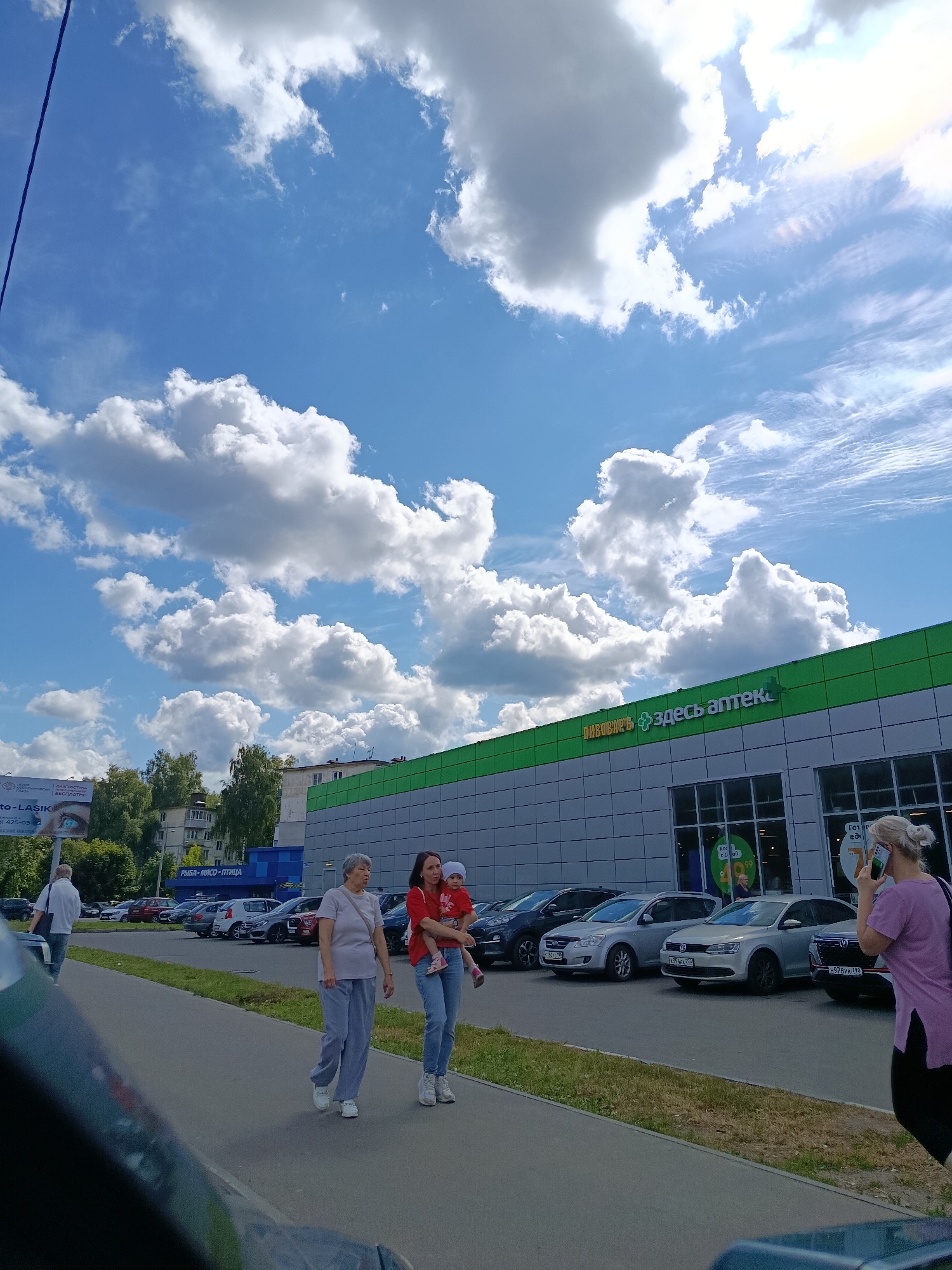 In Orekhovo-Zuyevo, Russia, on a sunny day with a bright blue sky dotted with white clouds, people walk along a paved sidewalk in front of a modern building with a green and grey facade. The building has large windows, and a prominent green sign reads "ЗДЕСЬ АПТЕК" which translates to "PHARMACY HERE". Arow of cars is parked in front of the building. On the sidewalk, an older woman in grey sweatpants and a light grey t-shirt walks to the left, while a younger woman in a red t-shirt and blue jeans holds a small child in her arms. To the far right, a woman with blonde hair tied in a bun is walking away from the camera, looking at her phone. In the background, trees and apartment buildings are visible. A billboard is also present on the left side of the image.