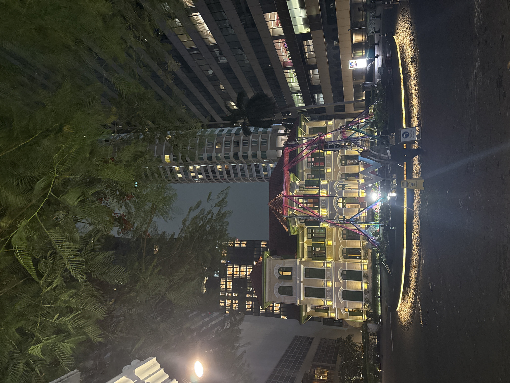 The image depicts an outdoor scene at night, likely in Bangkok, Thailand. In the foreground, lush green foliage from trees partially obscures the view. Behind the trees, a brightly lit, multi-story building with arched windows and decorative architectural elements stands out against the dark sky. The building is illuminated with warm yellow lights, highlighting its facade.
To the left of this building, a modern, dark glass skyscraper rises, its windows aglow with interior lights, suggesting it's a commercial or residential complex. Connecting the two buildings, or perhaps in front of them, is a colorful, illuminated structure that resembles part of an amusement ride or a decorative art installation. It features a series of colorful beams and lights.
The ground in the right portion of the image is paved, with a curved, illuminated pathway made of stones and a yellow light strip. A parking sign is visible on a post, indicating a designated parking area. The overall ambiance is one of a lively urban night scene, possibly a commercial district, entertainment venue, or a well-designed public space. The time of day is clearly night, and the weather appears clear. There are no people clearly visible in the image, but the illuminated buildings and pathway suggest a place that is open and active.