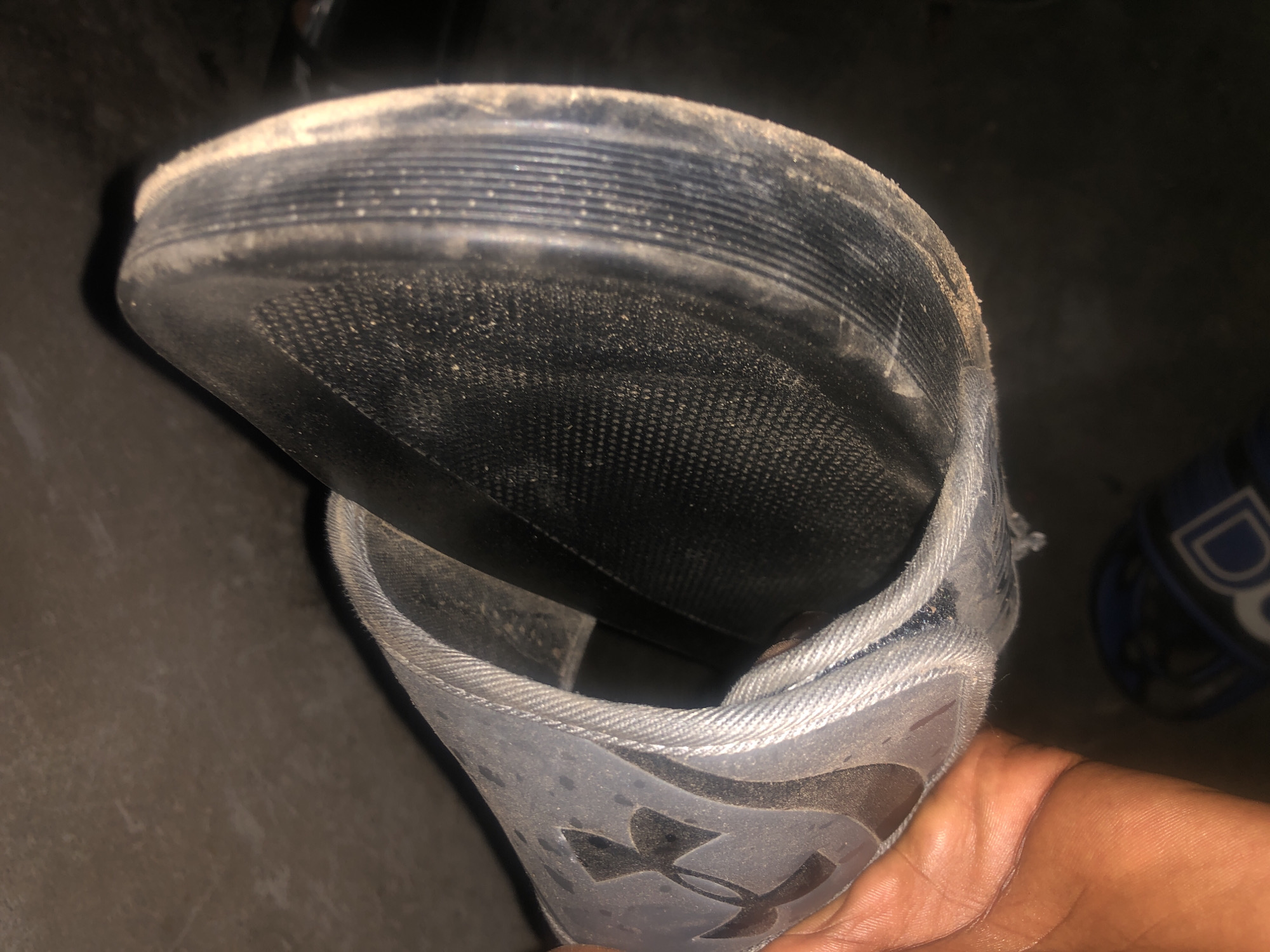 A close-up, low-angle shot shows a dirty, light-grey Under Armour slide sandal held by a person's hand. The sandal is positioned horizontally, with the sole facing the viewer. The sole has a textured, black surface with a repeating pattern of small dots, and the outer edge is black with ribbed detailing. The strap of the sandal is a light grey fabric with a visible seam and the Under Armour logo embossed on the upper side. The sandal appears to be worn and dusty. In the background, a dark, textured surface is visible, possibly a floor or wall. Below the sandal, another sandal with blue and white markings is partially visible. The scene is dimly lit, with shadows obscuring some details. The location is identified as Jalingo, Nigeria.