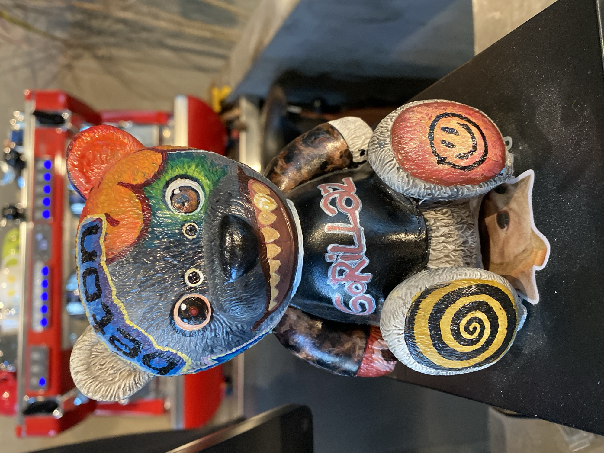 A custom-painted teddy bear sits on a black surface in front of a red espresso machine. The bear is black with a colorful, abstract design on its head, including blue, yellow, orange, and green sections. It has large, expressive eyes and an open mouth revealing jagged, yellow teeth. The bear wears a black t-shirt with the text "GORILLA" in red. Its paws are also painted: the left paw has a yellow and black spiral design, and the right paw has a black smiley face on a red background. The espresso machine in the background is red with silver accents and illuminated blue buttons. The scene appears to be indoors, possibly in a cafe or shop. There is no visible indication of its location in Da Nang, Vietnam.