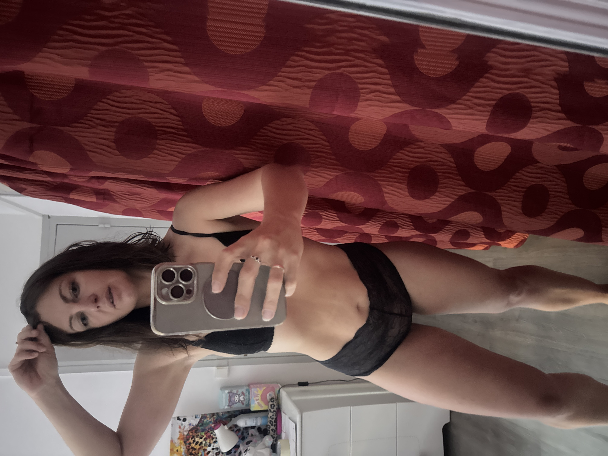 A woman is taking a mirror selfie in what appears to be a bedroom. She is wearing black lace lingerie and standing in front of a red patterned curtain. To her left, a white dresser is visible with various items on top, including what looks like a lamp and some decorative boxes. The flooring is a light-colored wood. The background is blurred, but suggests an indoor setting, possibly a residential room. There are no specific landmarks or indications of the city of Saint-Claude, France.