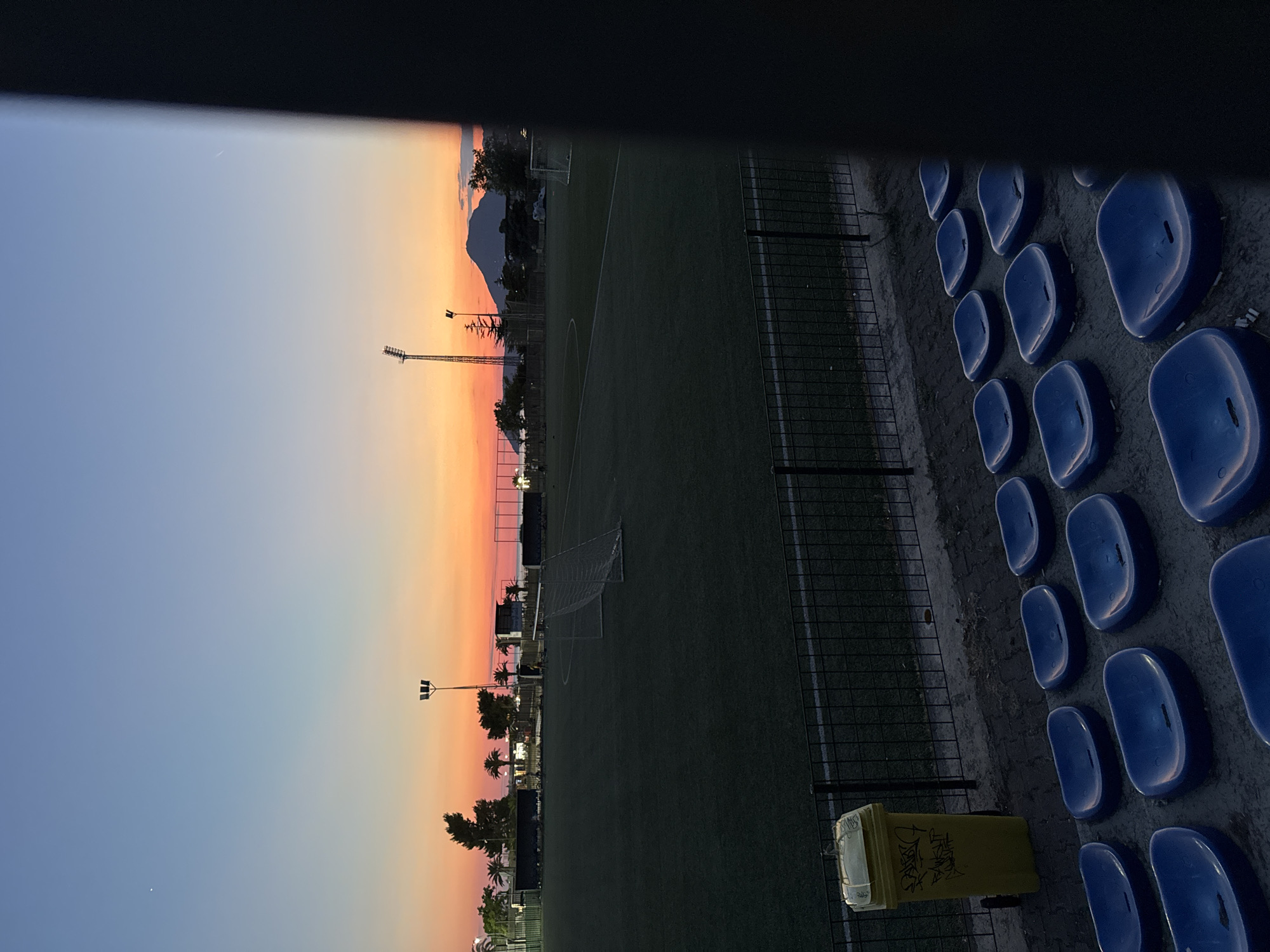 The image depicts a soccer field at sunset in Bar, Montenegro. The sky is a gradient of blue and orange, with a faint star or planet visible in the upper left corner. A single light pole with multiple lamps stands near the horizon, casting a faint glow. In the distance, a mountain range is silhouetted against the colorful sky. The soccer field itself is covered in green turf, with a net in the center and lines marking the boundaries. In the foreground, there are rows of empty blue stadium seats, and a yellow trash can with graffiti is visible on the left side of the frame. A black fence separates the seating area from the field. The overall scene is quiet and serene, suggesting the end of a day.