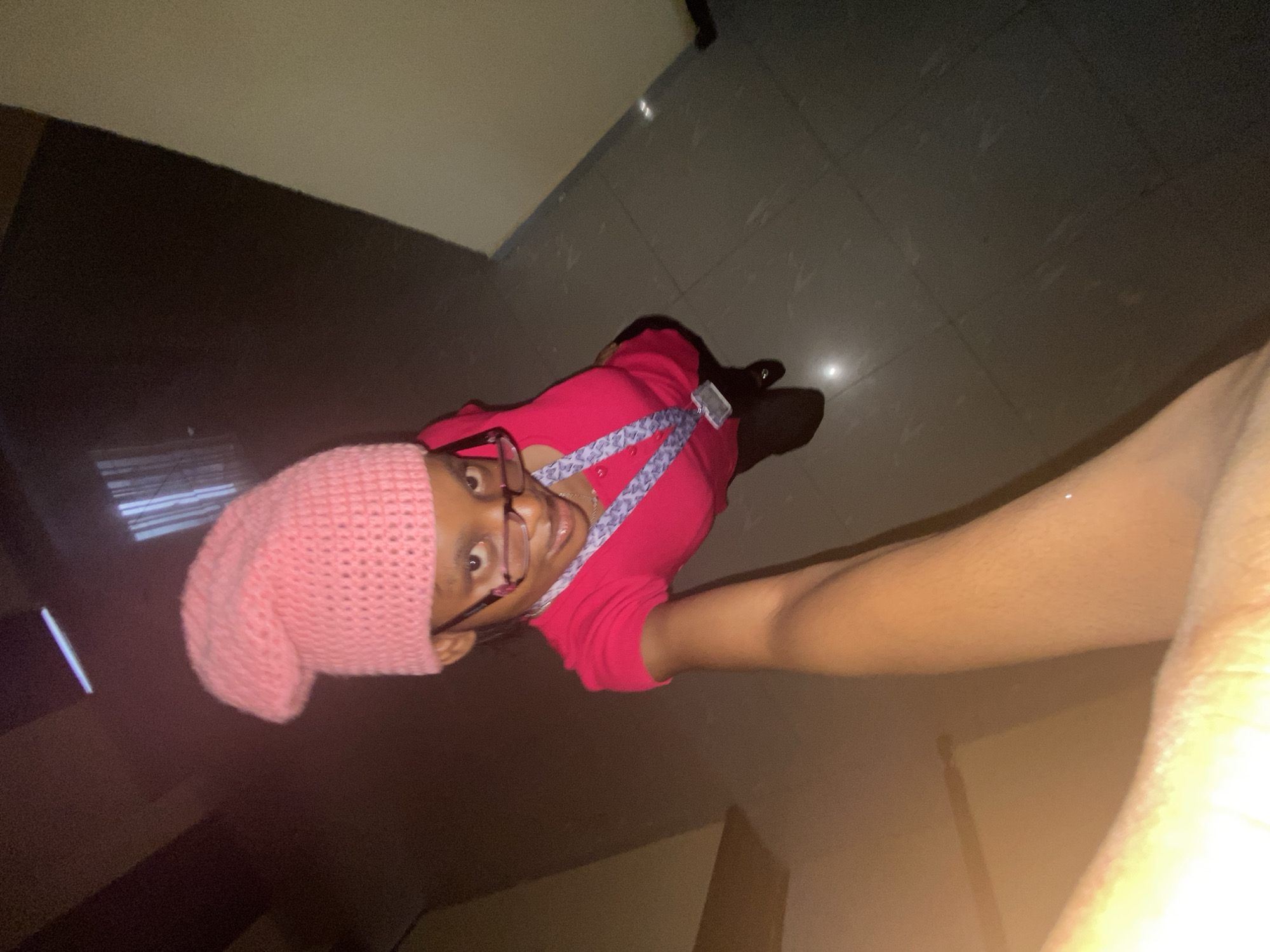A person is looking up at the camera. They are wearing a pink knit hat and glasses. They are also wearing a pink shirt with a purple and white patterned lanyard. The person is standing on a tile floor. The walls are beige.