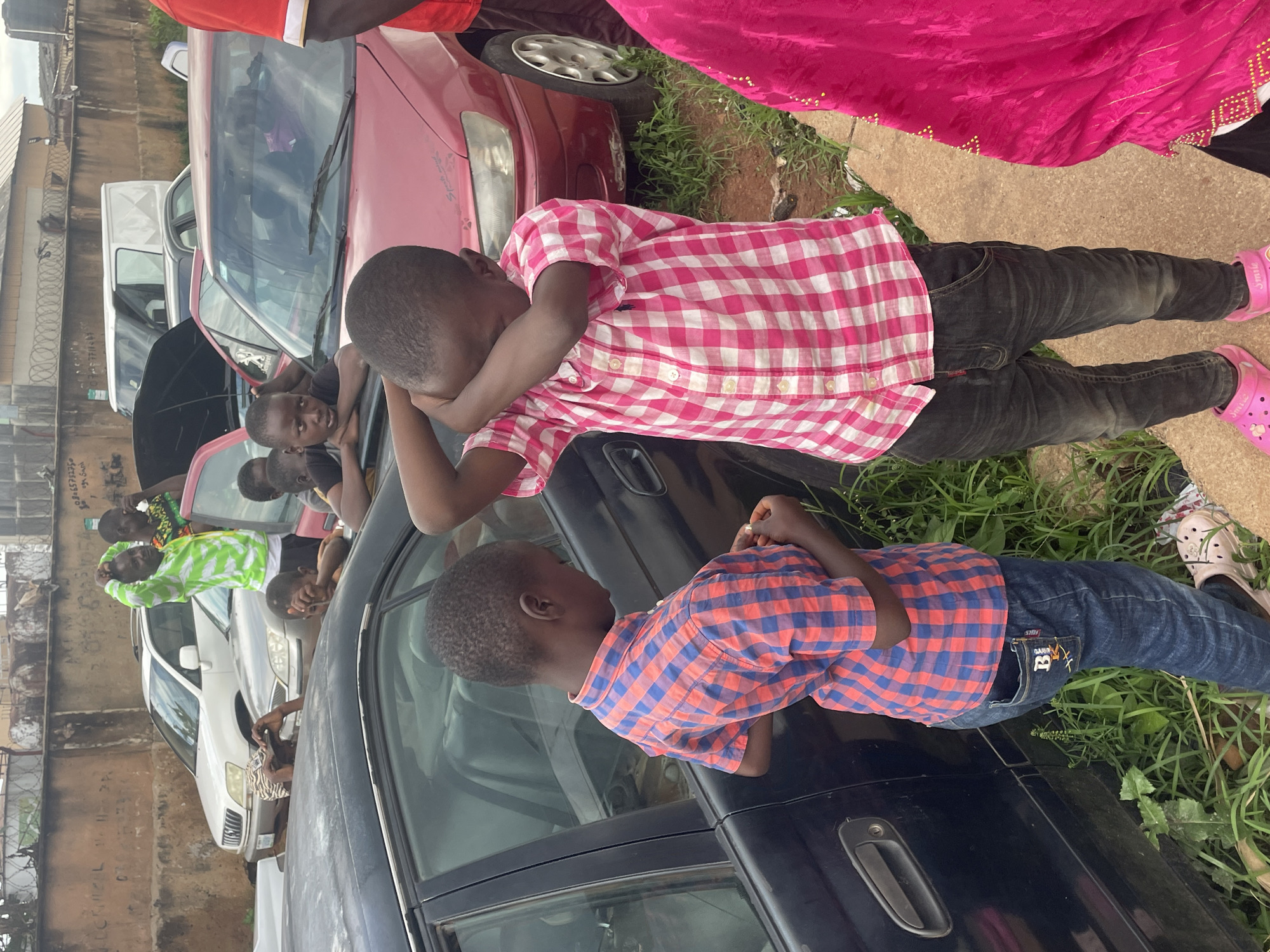 The image shows an outdoor scene in the city of Jos, Nigeria, characterized by a line of parked vehicles and a dirt and concrete walkway. The foreground features two young boys. The boy on the left, wearing a red and blue plaid shirt and blue jeans, stands looking down at his hands. The boy on the right, in a pink and white plaid shirt and dark jeans, has his arms covering his face. To the right of the boys, a person in a bright pink garment is partially visible.
In the background, several cars are parked, including a dark-colored sedan to the left, a red sedan in the center, and a white van and other vehicles further back. The area appears to be a residential or informal commercial space, with a brick wall and some vegetation visible. The lighting suggests it is daytime, possibly overcast.