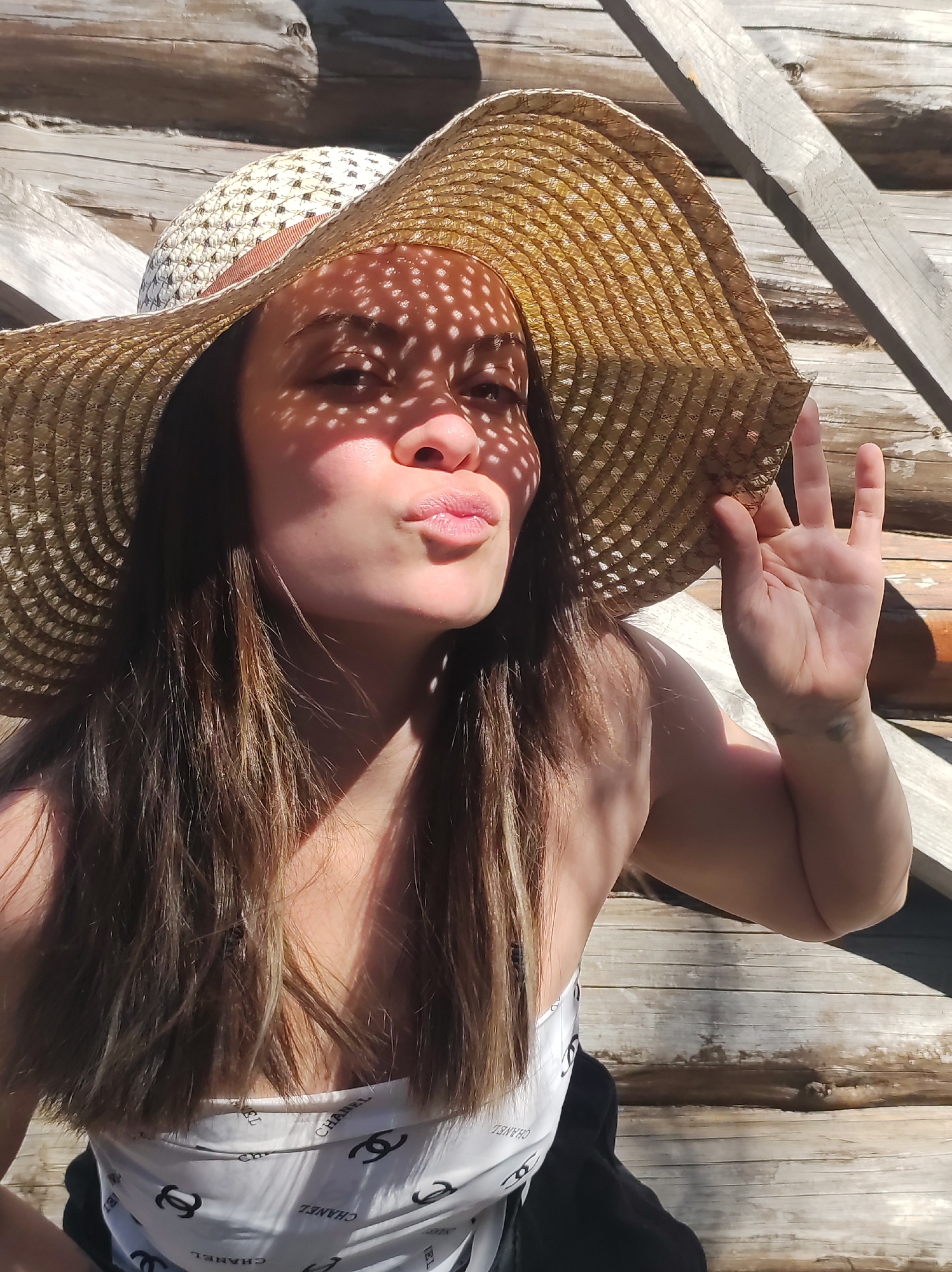 A young woman is captured in a sunlit, close-up portrait, likely a selfie, against a rustic wooden background. She appears to be in her 20s or 30s, with long, straight dark brown hair falling over her shoulders. She wears a wide-brimmed, woven straw hat, the open weave of which casts distinctive dappled circular shadows across her forehead, eyes, and nose.
Her expression is playful, with her lips puckered into a "kissy face." Her left hand is raised near the brim of her hat, forming a gesture that resembles an "okay" sign or a three-finger salute, with her palm facing forward. She is wearing a white strapless or off-the-shoulder top adorned with repeated black "CHANEL" logos.
The background consists of weathered, light-colored wooden logs or planks, suggesting an outdoor setting like a cabin or a wooden fence, consistent with a location in Фоки, Russia. Strong, defined shadows are cast across the scene by the bright sun, indicating a clear, sunny day, likely midday or early afternoon. The overall mood conveyed is casual and cheerful.