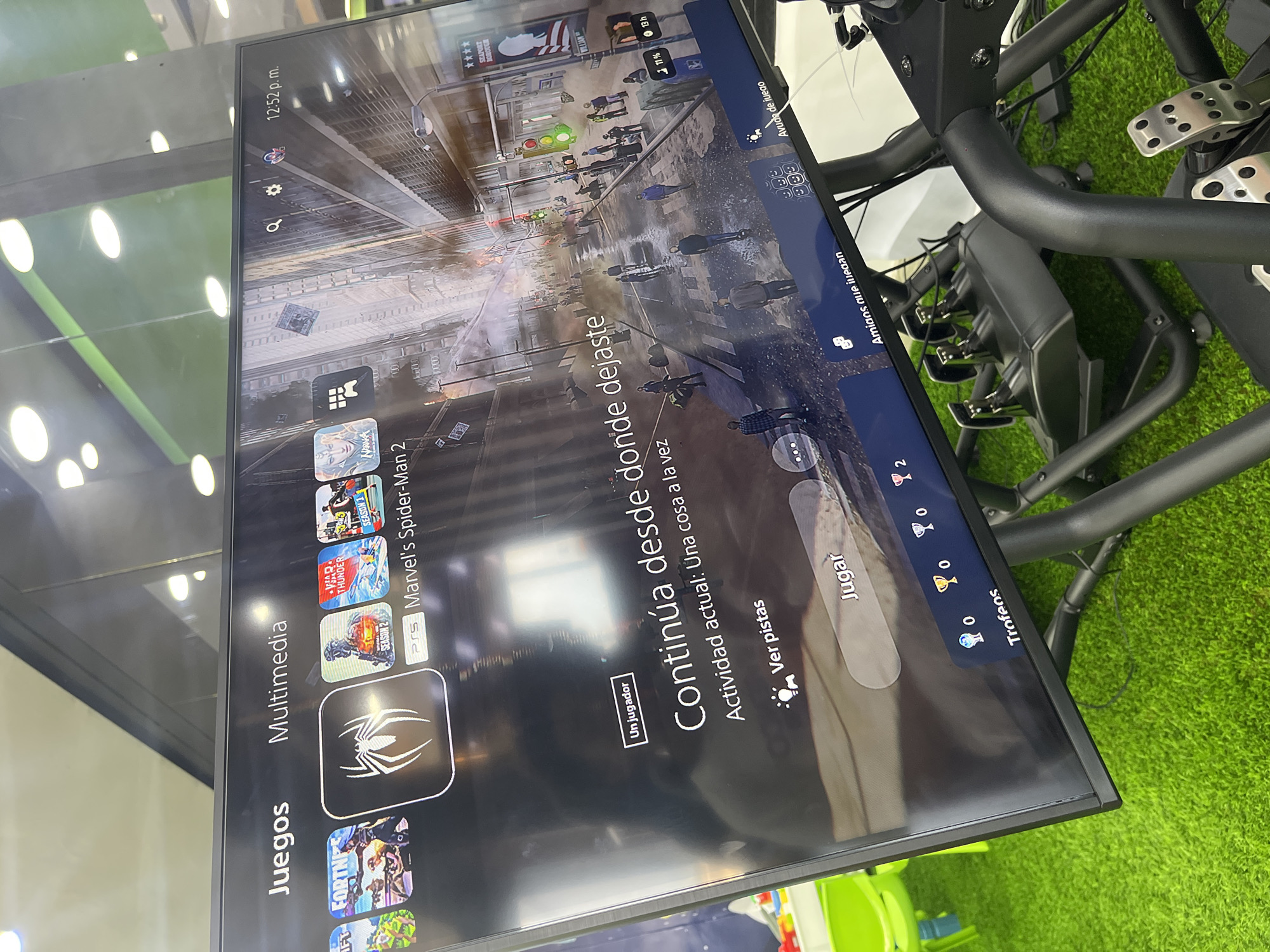 The image shows a PlayStation 5 screen displaying the home menu in Spanish. The user is currently highlighted on "Marvel's Spider-Man 2" under the "Multimedia" and "Juegos" (Games) sections. The highlighted game has a "Continue" option displayed. The screen also shows the system's trophy count (zero in all categories), online friends playing, and the current time (12:52 p.m.). The screen is reflecting light from the surrounding environment. In the background, a steering wheel and pedal set for racing games can be seen, as well as some green turf. The image does not suggest any connection to the city of Cotoperíz in Venezuela.