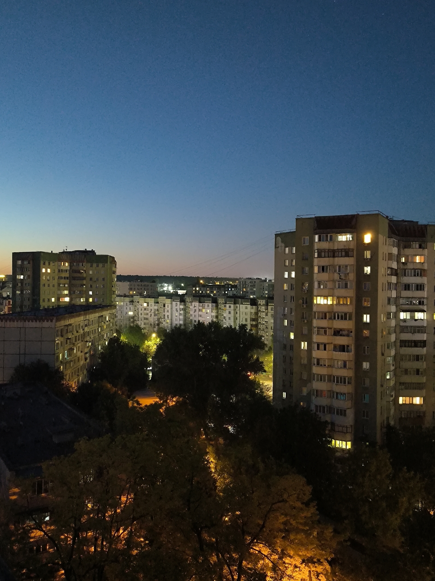 The image depicts an urban twilight scene in Chișinău, Moldova, featuring a panorama of Soviet-era apartment blocks against a darkening sky. The primary subject is a cluster of multi-story residential buildings, characterized by their utilitarian design and weathered facades. Many windows are illuminated from within, casting a warm glow that contrasts with the cool blues and purples of the evening sky. In the foreground, dark silhouettes of trees with visible foliage suggest a residential area with green spaces. The overall impression is of a quiet evening in a densely populated urban environment. There are no discernible people or specific actions taking place.