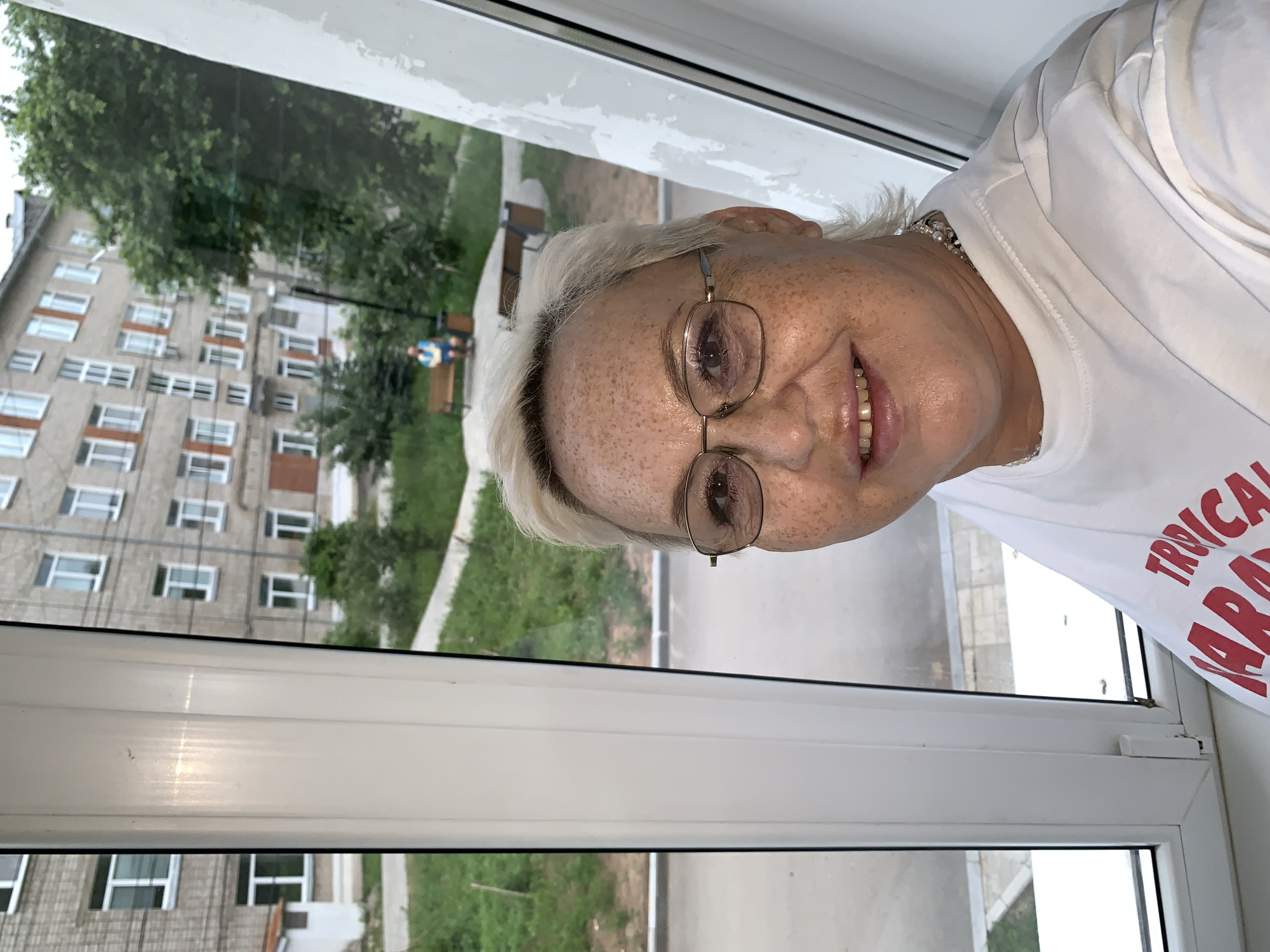 A woman with short, blonde hair and freckles, wearing glasses and a white t-shirt with red lettering that says "TROPICAL PARADISE" is taking a selfie indoors, with a window to her left. Outside the window, a building with multiple floors and windows is visible, along with some trees and a person sitting on a bench. The building appears to be an apartment complex in Chaikovsky, Russia, judging by the architectural style which is common in Soviet-era residential areas. The scene suggests a residential setting within the city.