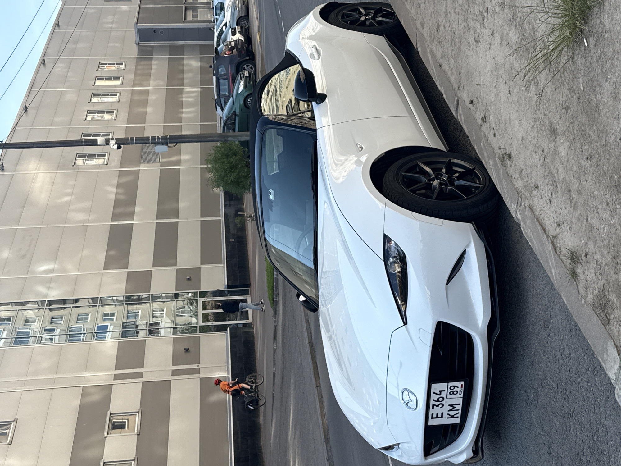 The image captures a white Mazda MX-5 Miata convertible parked on a city street in Saint Petersburg, Russia. The car is facing forward, with its front license plate visible, reading "E 364 KM 89" and featuring the "RUS" designation. The car is parked next to a concrete curb, with a paved street in front of it.
In the background, a large, modern building with a grey and beige striped facade dominates the scene. The building has numerous windows, and reflections of the sky and surrounding environment are visible on its surface. A streetlamp with a security camera is positioned near the building.
Further in the background, several other cars are parked along the street. To the left of the building, a person wearing an orange jacket and a red helmet is riding a bicycle on a sidewalk. Another person in dark clothing is walking on the sidewalk near the entrance of the building. The sky is clear and appears to be daytime. The overall environment suggests an urban setting.