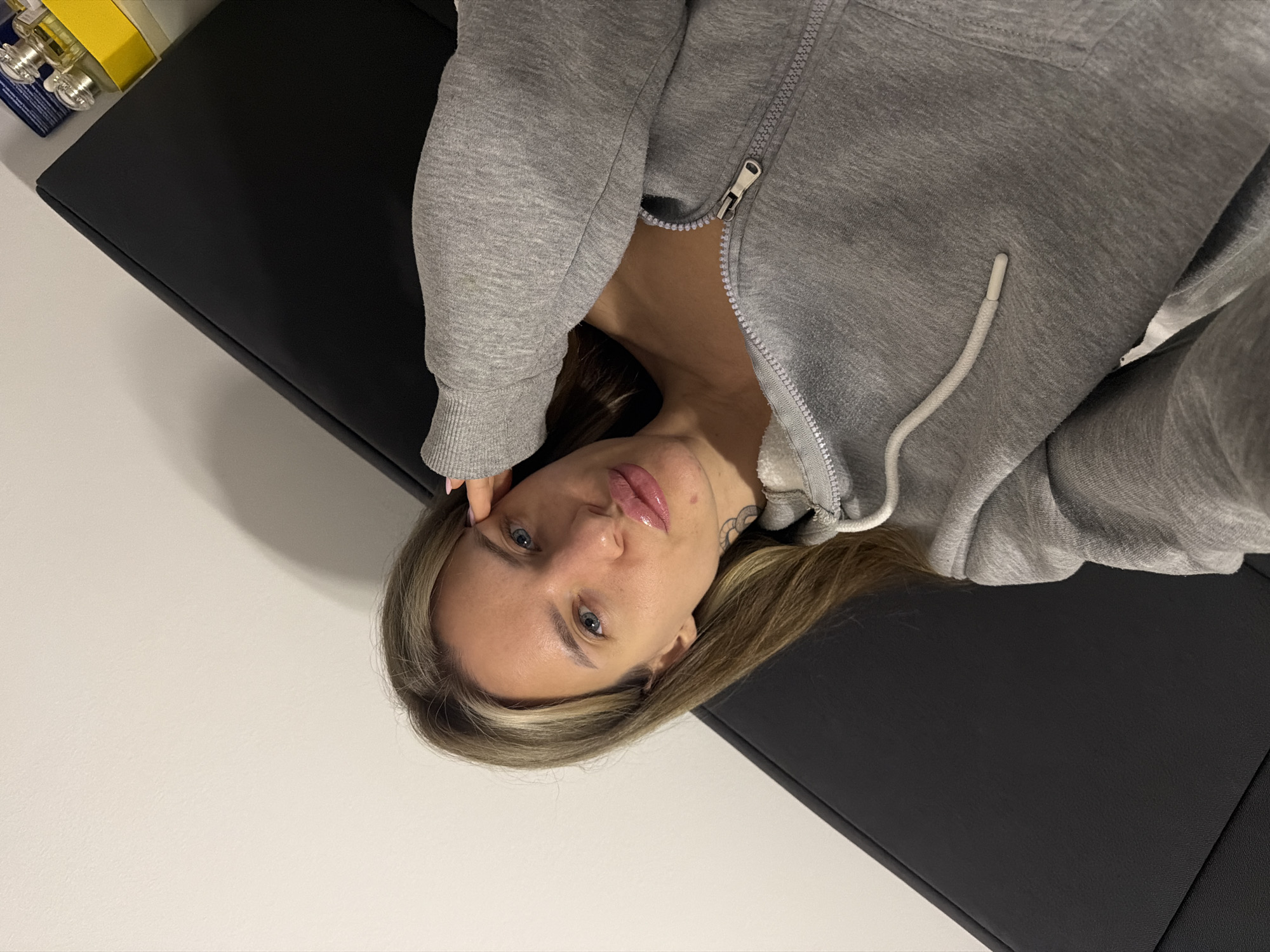 A woman lies on a black examination table, wearing a gray zip-up hoodie. Part of a white wall is visible, as well as what appears to be medication or cleaning supplies on a surface beside the table. It is not possible to verify the location as Berlin, Germany, from this image.