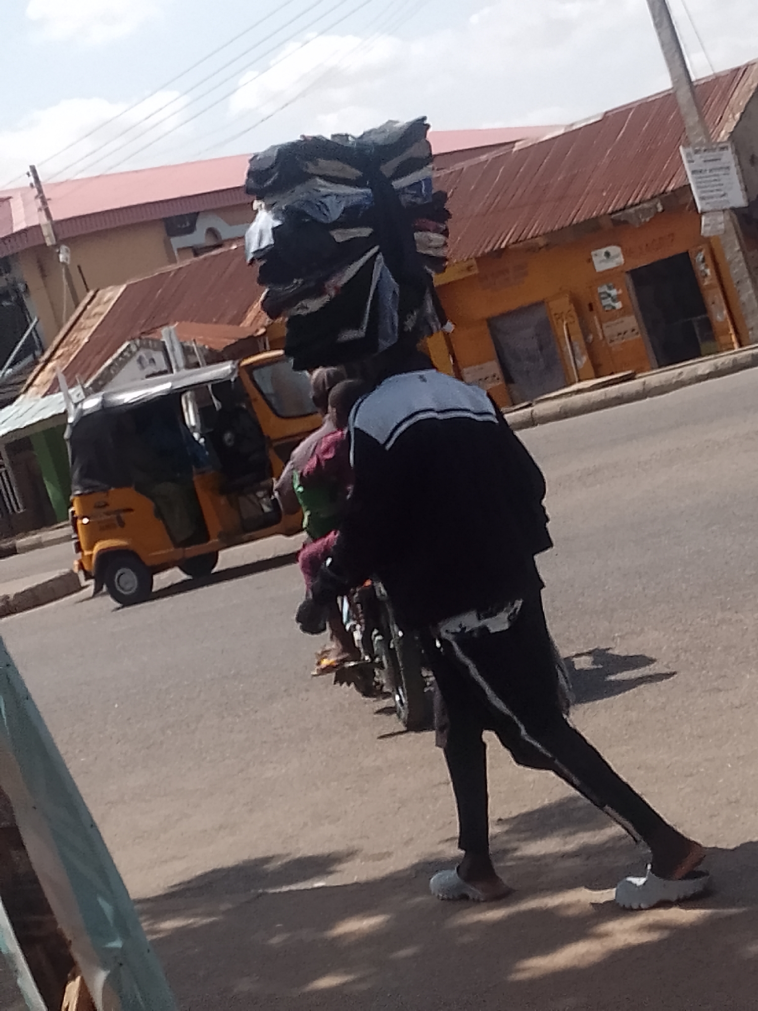 This image captures a street scene in Pantame, Nigeria, under bright daylight. The central subject is a person, likely a street vendor or hawker, walking with a large, stacked bundle of what appears to be clothing or textiles balanced on their head. They are wearing a black jacket with white accents on the shoulders and dark trousers, paired with light-colored sandals. The person is walking on the side of a paved road, next to a motorcycle which has a passenger clinging to the driver's back. Long shadows are cast on the ground, indicating the sun is overhead, possibly mid-morning or afternoon.
In the background, a yellow auto-rickshaw (keke Napep) is visible on the road, either parked or moving slowly. Beyond the road, there are buildings with reddish-brown and orange corrugated iron roofs. One prominent building is painted orange/yellow with closed doors or windows, suggesting a shop or commercial establishment. Power lines crisscross the bright, partly cloudy sky. To the far left, a blurred green object suggests another element in the immediate foreground, possibly part of another vehicle or a barrier. No legible text is visible on signs or vehicles in the image.