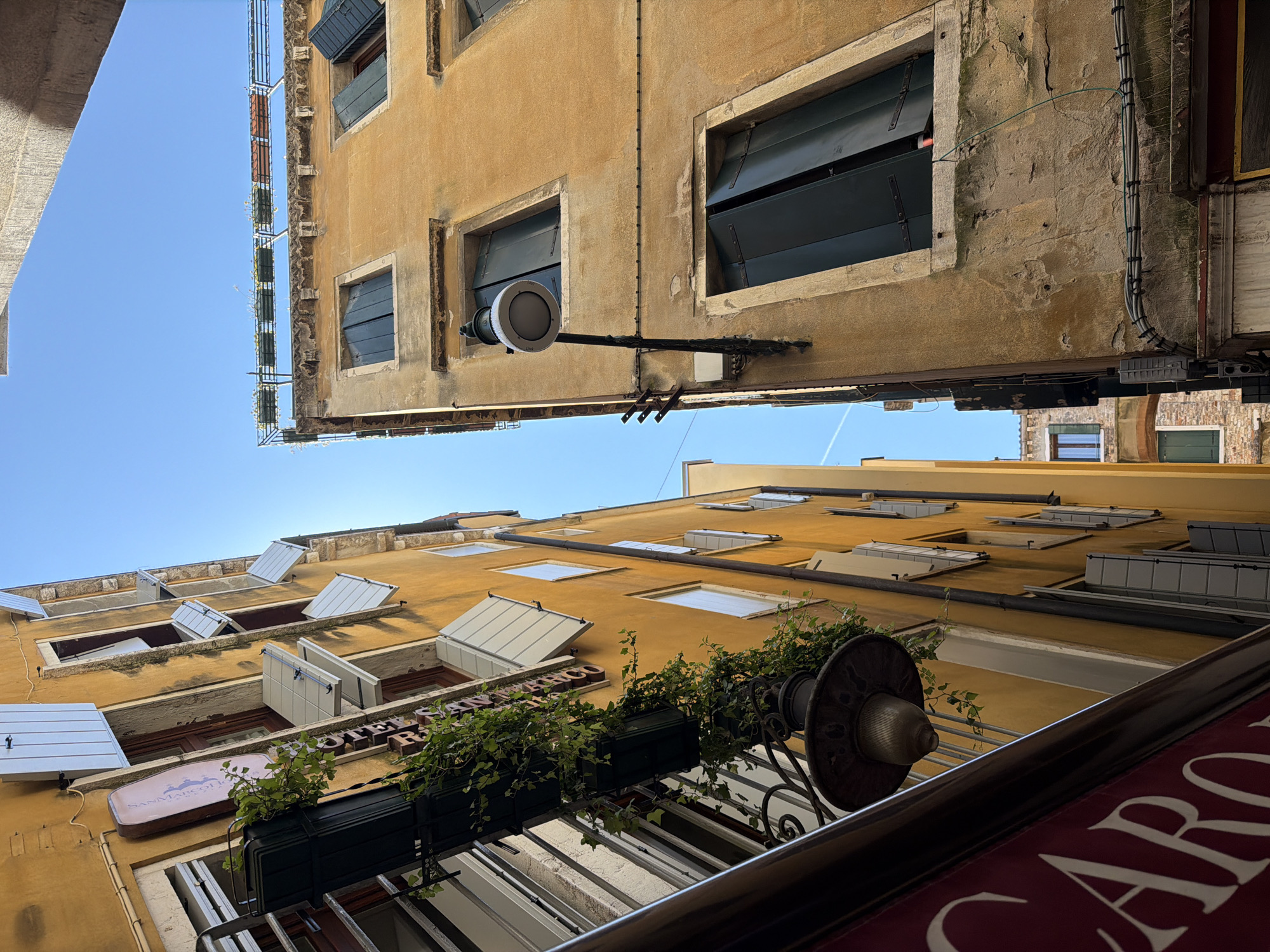 This media file is a low-angle shot looking up at the facades of buildings in Venice, Italy. The sky above is a clear, bright blue, suggesting a sunny day. The architecture is characteristic of Venice, with narrow buildings constructed of aged, yellow stucco.
The upper portion of the image shows a building with several windows, some of which have dark green shutters partially open. A utilitarian light fixture with a ribbed glass shade is mounted on the wall between two windows. Wires and conduit run along the wall, indicating modern utilities on an older structure.
The lower portion of the image focuses on another building facade, also painted in a warm yellow hue. This building has numerous windows, many of which are fitted with angled, white awnings or shutters, some of which are open. A dark green planter box filled with lush green ivy hangs from a railing, adding a touch of nature to the urban environment. A vintage-style street lamp with a ribbed glass shade is also visible, attached to the same railing.
Visible text includes the partial word "CARO" in white on a dark red banner or awning in the foreground, suggesting a business or establishment. Another sign above the planter box reads "SAN MARCO..." in a stylized font. The overall impression is one of exploring the charming, narrow alleyways of Venice, where historic buildings are interspersed with touches of greenery and signs of daily life. There are no people visible in the image. The lighting suggests it is daytime, likely late morning or early afternoon.