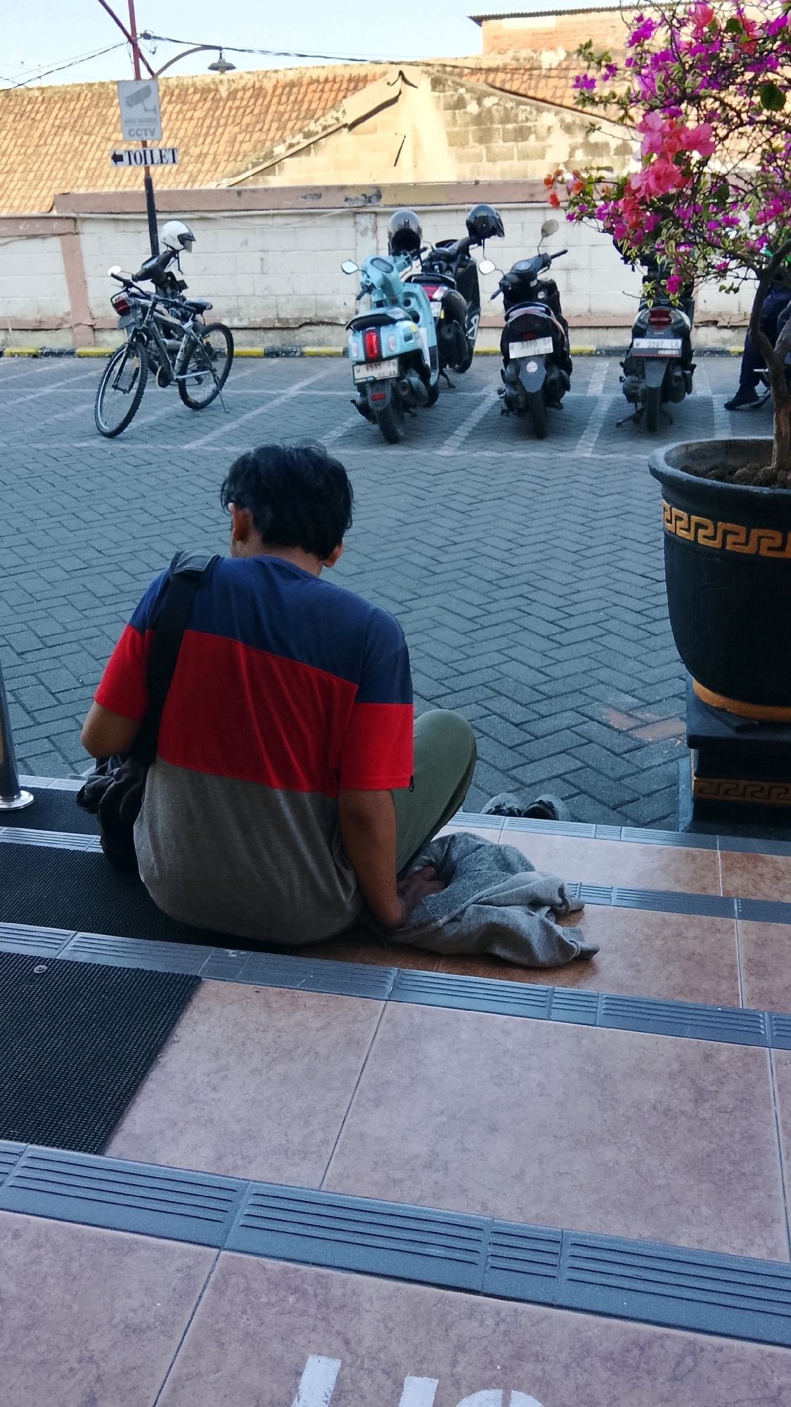 A rear view of a man sitting on the stairs facing away from the camera in Ngipik, Indonesia. He is wearing a t-shirt with horizontal bands of red, blue, and grey, and green pants. A dark bag is slung over his left shoulder. Beside him, a grey cloth is draped. The stairs are tiled with light brown and grey-brown tiles, with textured grey strips at the edges. Behind him, a paved parking area is visible with several motorbikes and a bicycle parked. A sign indicating "CCTV" and "TOILET" with an arrow is mounted on a pole. A tree with vibrant pink and purple flowers is to the right of the motorbikes, and a large black planter pot is also present. The background shows a building with a tiled roof.
