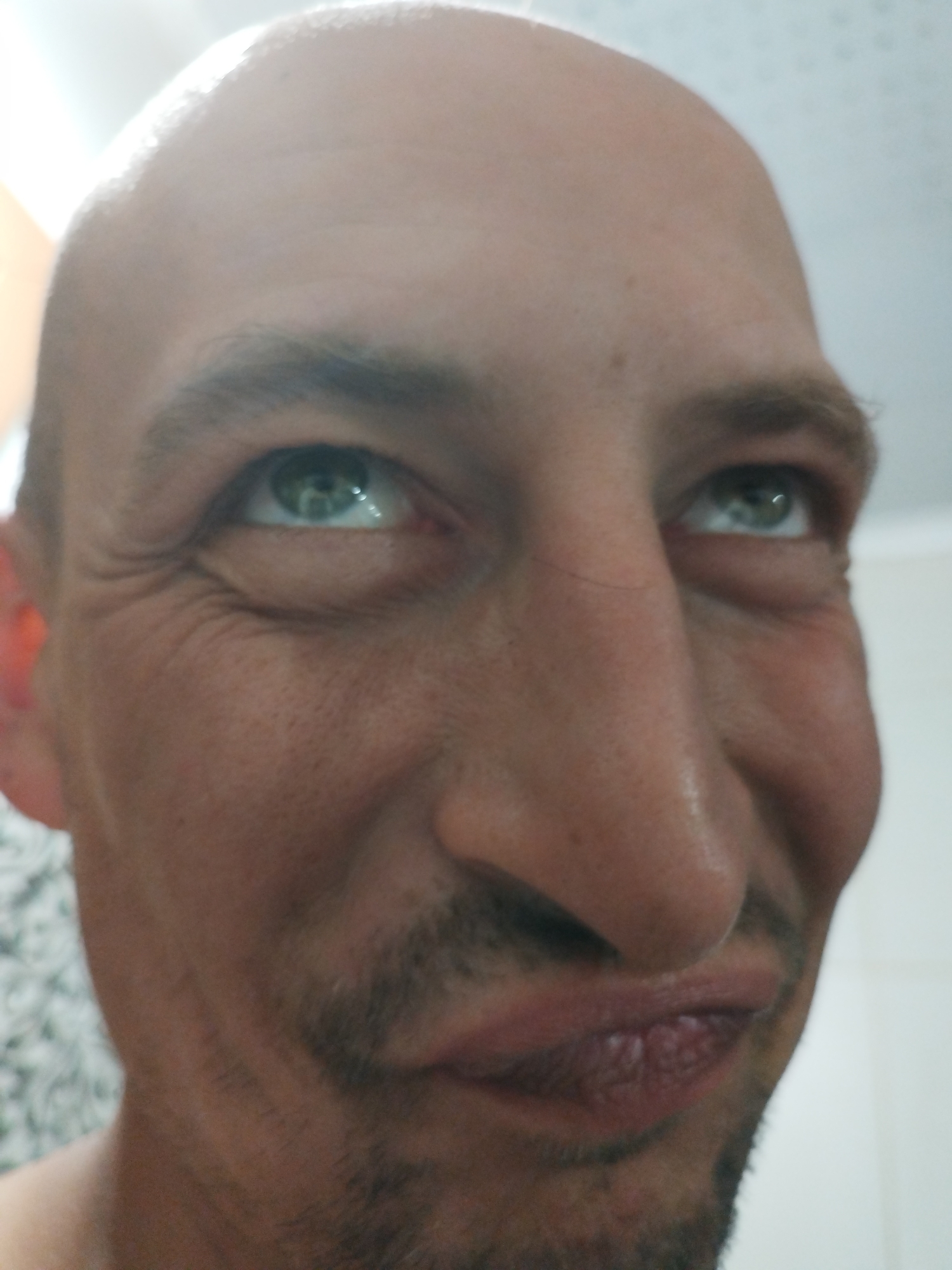 A close-up, low-angle, candid shot shows the face of a bald man from the nose up. His eyes are looking upwards and to the left, and he has a subtle grimace. The lighting is bright and seems to be coming from above, casting shadows on his face, particularly under his nose and chin. The background is out of focus and appears to be an indoor setting with a light-colored wall and a patterned surface on the left.