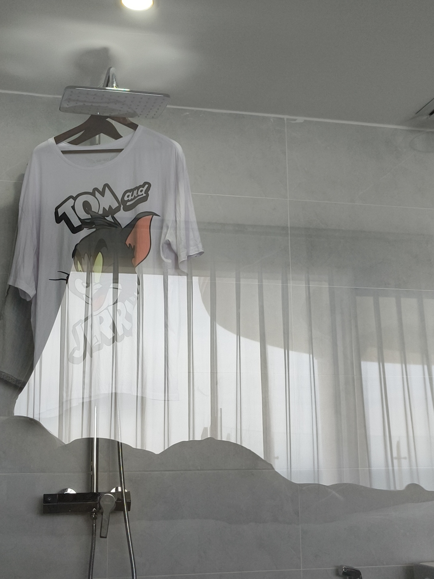 A white t-shirt with a graphic of Tom and Jerry is hung on a hanger from a showerhead in a tiled bathroom, possibly in Phu Thuy Ward, Vietnam. The t-shirt is draped over the hanger and the showerhead extends from the ceiling. The walls are covered in gray tiles. The image appears to be taken through a glass surface, as evidenced by reflections of sheer curtains and possibly a window or balcony. A shower faucet is visible on the tiled wall below the t-shirt. The scene suggests an attempt to dry the t-shirt by utilizing the bathroom's ventilation or warmth, or possibly as a temporary measure.