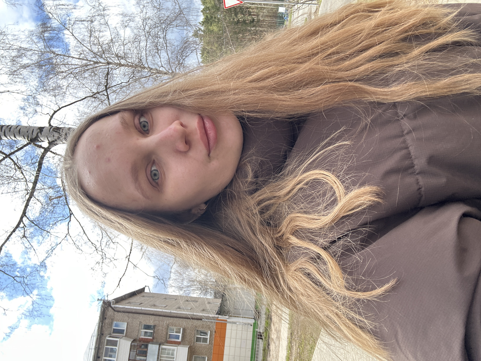 A close-up selfie captured outdoors features a young woman with long, wavy blonde hair and light blue-green eyes. She has a neutral expression, with slightly parted lips. Her skin appears fair, and she has a few visible blemishes on her forehead and nose. She is wearing a dark brown, possibly padded or quilted, jacket. The photo is taken from a low angle, looking upwards.
The background reveals a clear sky with scattered white clouds and bare tree branches, suggesting either late winter or early spring. A prominent birch tree with its distinctive white bark is visible on the left side of the frame. In the distance, parts of a multi-story, concrete apartment building with several windows and balconies are visible. There are also hints of other buildings or structures, and what appears to be a road or path. A red triangular road sign is partially visible in the upper right background. The overall impression is that of an urban or suburban setting, consistent with Chaikovsky, Russia, during a cooler season.