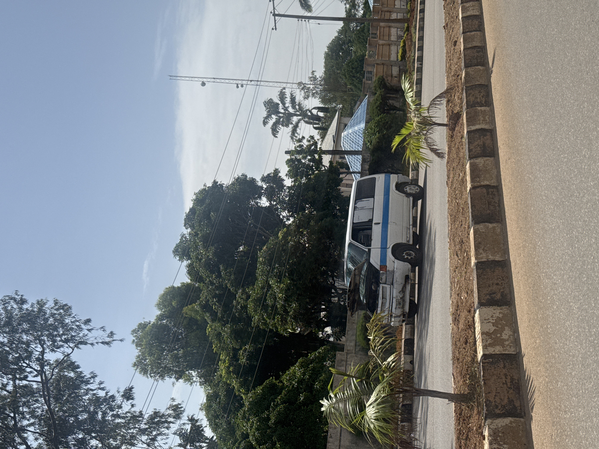 The image depicts a street scene in Bar Jirgi Summa, Nigeria. The perspective is angled, giving a sense of looking down the road. The foreground is dominated by an asphalt road with a textured, stone curb along the right side. Some sparse, dry grass grows between the curb stones.
A white van with a blue stripe is parked on the left side of the road. The van appears slightly worn, with some visible rust and dirt. Behind the van, a variety of lush green trees and vegetation are prominent, creating a dense foliage backdrop. One particularly tall palm tree with distinctive fronds stands out amongst the other trees.
In the background, there are buildings with what appears to be a blue-tiled roof. Further back, power lines and utility poles crisscross the sky. The sky itself is a clear, bright blue with some wispy white clouds, suggesting a sunny day. The overall impression is of a warm, possibly tropical, climate. The lighting indicates it is likely daytime, with sunlight casting shadows. No people are visible in the frame. There is no discernible text within the image. The scene appears quiet and static, with no activity explicitly taking place other than the presence of the parked vehicle and the natural elements of the environment.