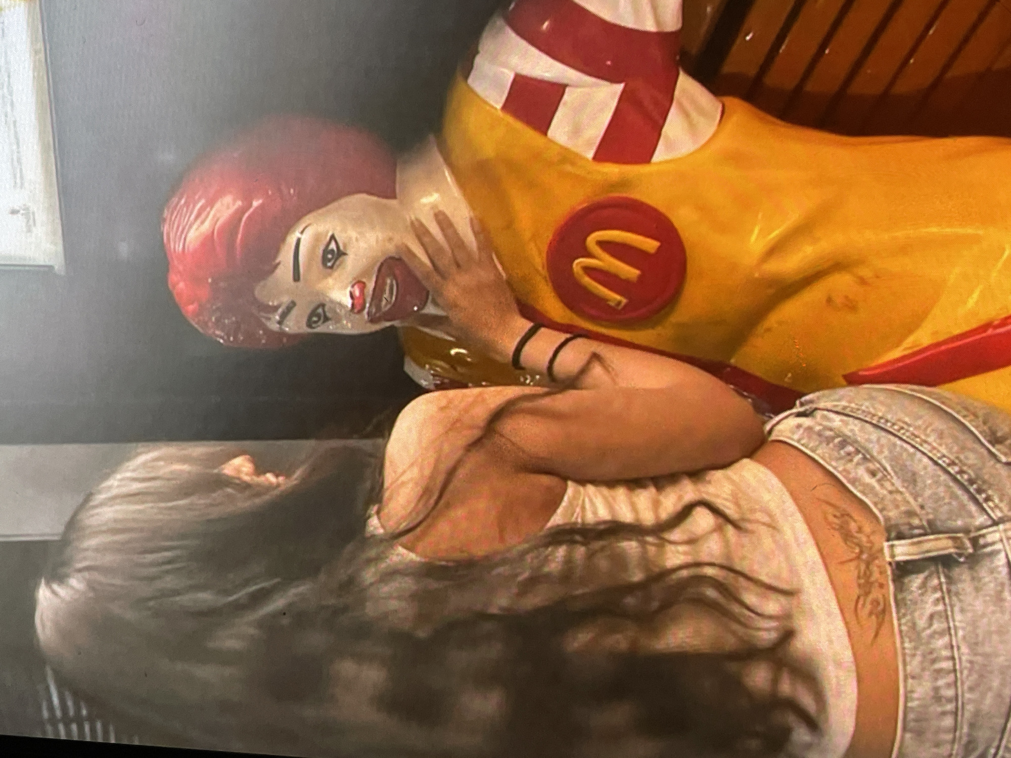 A woman with long, dark hair, wearing a white tank top and light-wash denim shorts, is lying down next to a Ronald McDonald statue in Harmanli, Bulgaria. The statue is predominantly yellow with red and white striped accents on the sleeves and collar. It features the iconic McDonald's logo on its chest. The woman's arm is resting on the statue. She has a tattoo on her lower back. The environment appears to be indoors, possibly in a McDonald's restaurant.