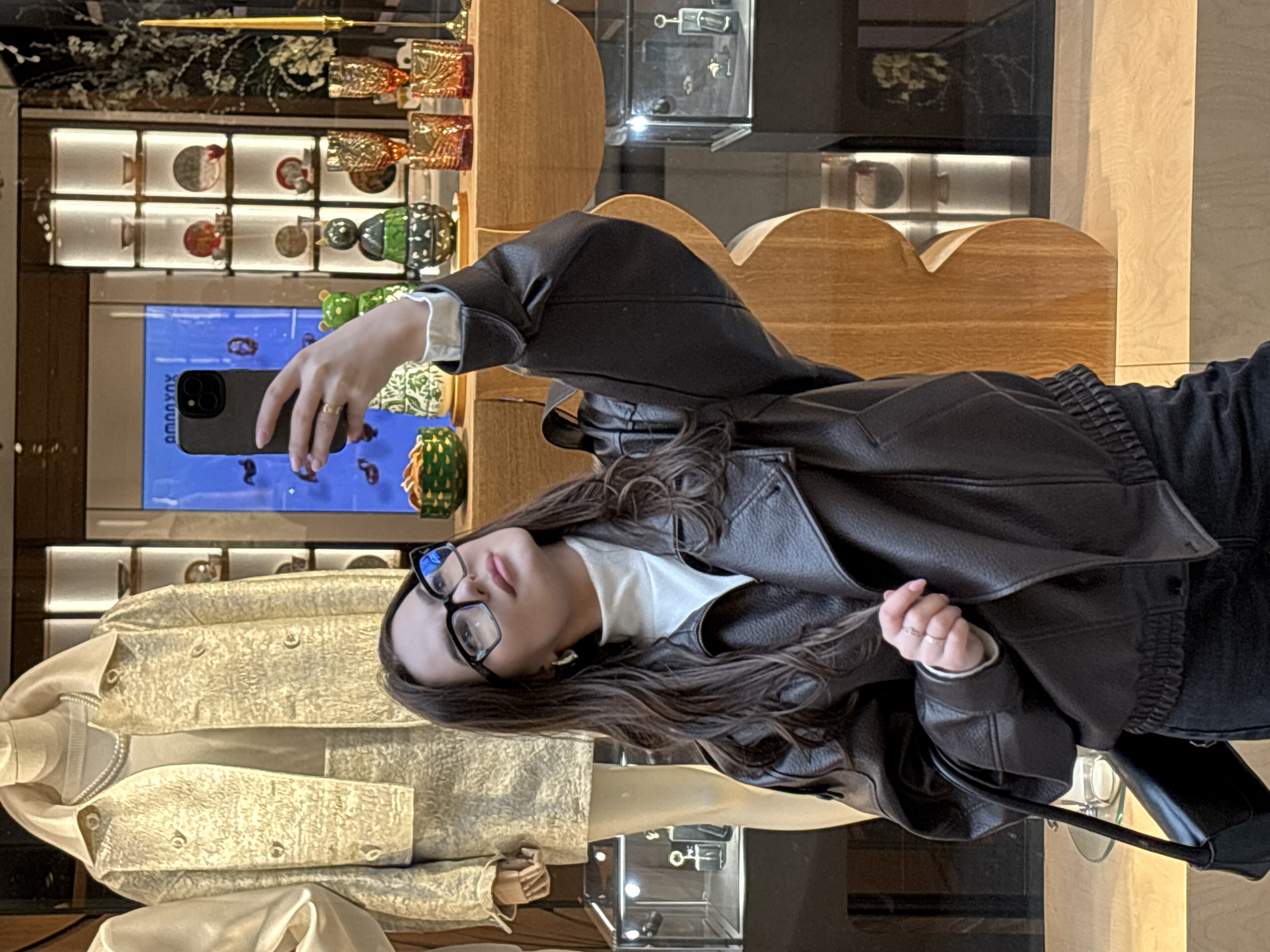 A woman wearing a brown leather jacket and glasses is taking a selfie with a smartphone inside a store. Behind her is a display case with various decorative items and a mannequin wearing a light-colored jacket. The store appears to be located in Moscow, Russia, based on the Cyrillic script on a screen in the display case.