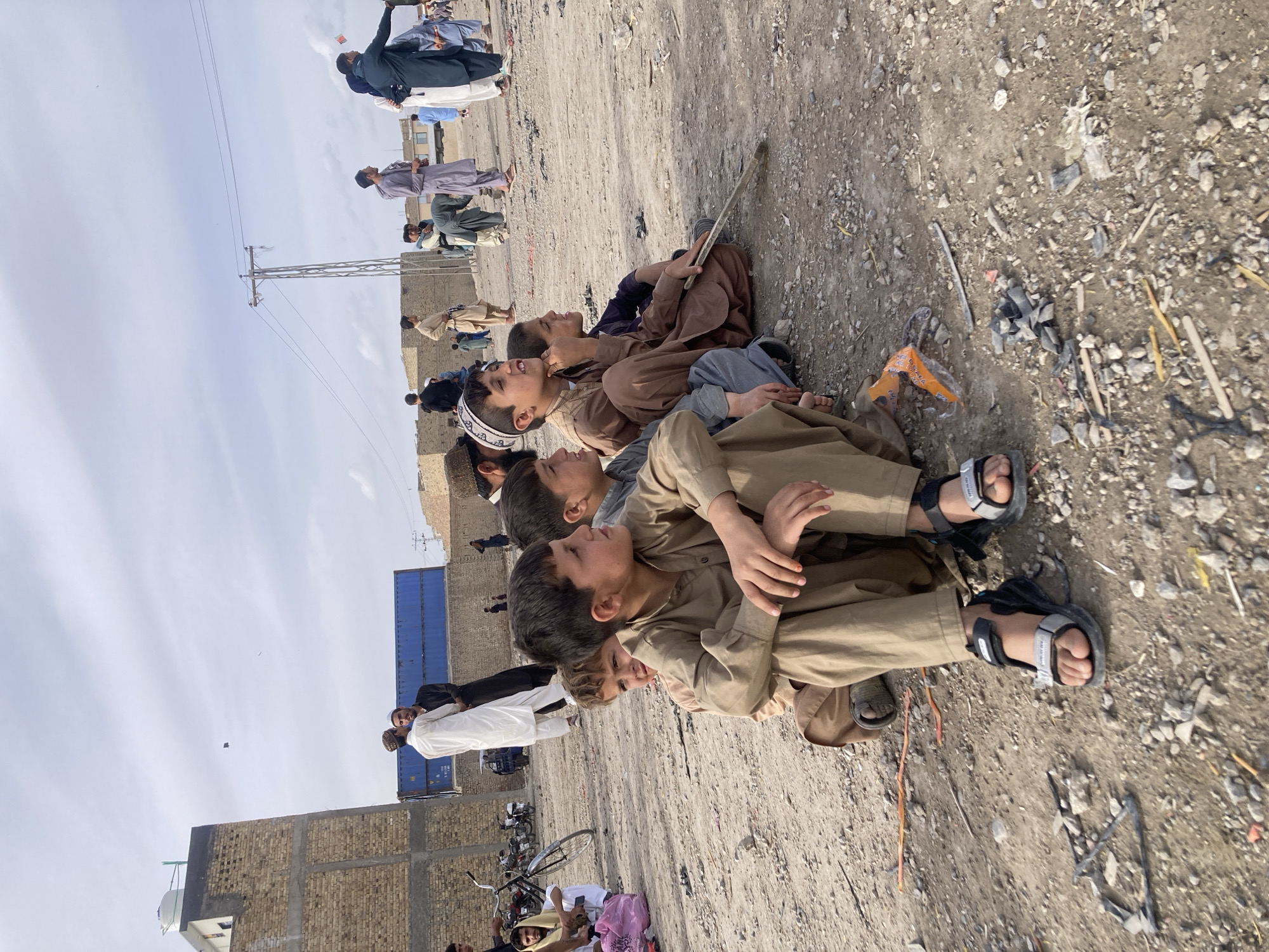 The scene depicts an outdoor gathering in Quetta City Tehsil, Pakistan, on a dusty, gravelly ground surface with scattered refuse. In the foreground, five young boys are seated in a row on the ground, facing right and looking upwards. They wear traditional shalwar kameez in shades of tan, brown, and grey. The boy closest to the camera wears black and grey sandals. Another boy further back wears a white skullcap. A boy in the middle holds a small stick.
In the mid-ground and background, numerous adult males are standing and walking. They are dressed in traditional South Asian attire, some with head coverings. Two men are conversing in the mid-ground: one in a white shalwar kameez and a white cap, the other in black attire. Behind them, two blue shipping containers are stacked. On the far left, bicycles and motorcycles are parked near a two-story brick building with an unplastered facade and a white water tank on its roof. On the right, a metal utility pole with power lines extends into the sky. Other distant brick structures and buildings are visible. The sky is partly cloudy, and at least two kites are observed flying; one is dark and diamond-shaped, the other smaller and lighter. A man in a blue shirt in the far background on the right appears to be involved with a kite or kite string. The ground is littered with small stones, plastic fragments, and other debris.