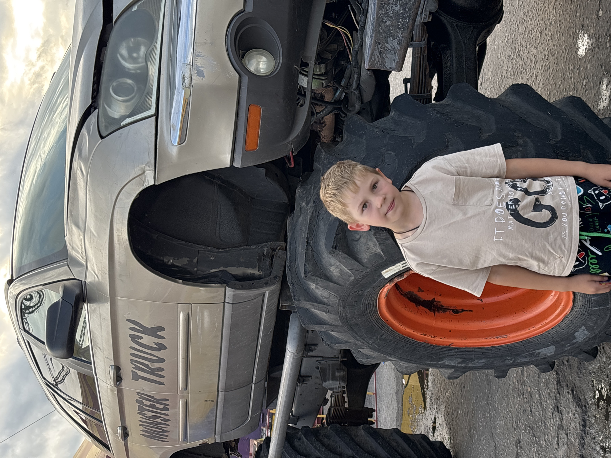 A young boy with blonde hair stands in front of a monster truck. The truck is a modified car, painted a light gold color with black lettering that reads "MONSTER TRUCK" on its side. The car is elevated on a heavy-duty chassis with enormous black tires that have bright orange rims. The boy is wearing a light tan t-shirt with a graphic print and patterned shorts. The background consists of an overcast sky and a paved surface that appears wet in places. The image was taken in the city of Korobovo, Russia.